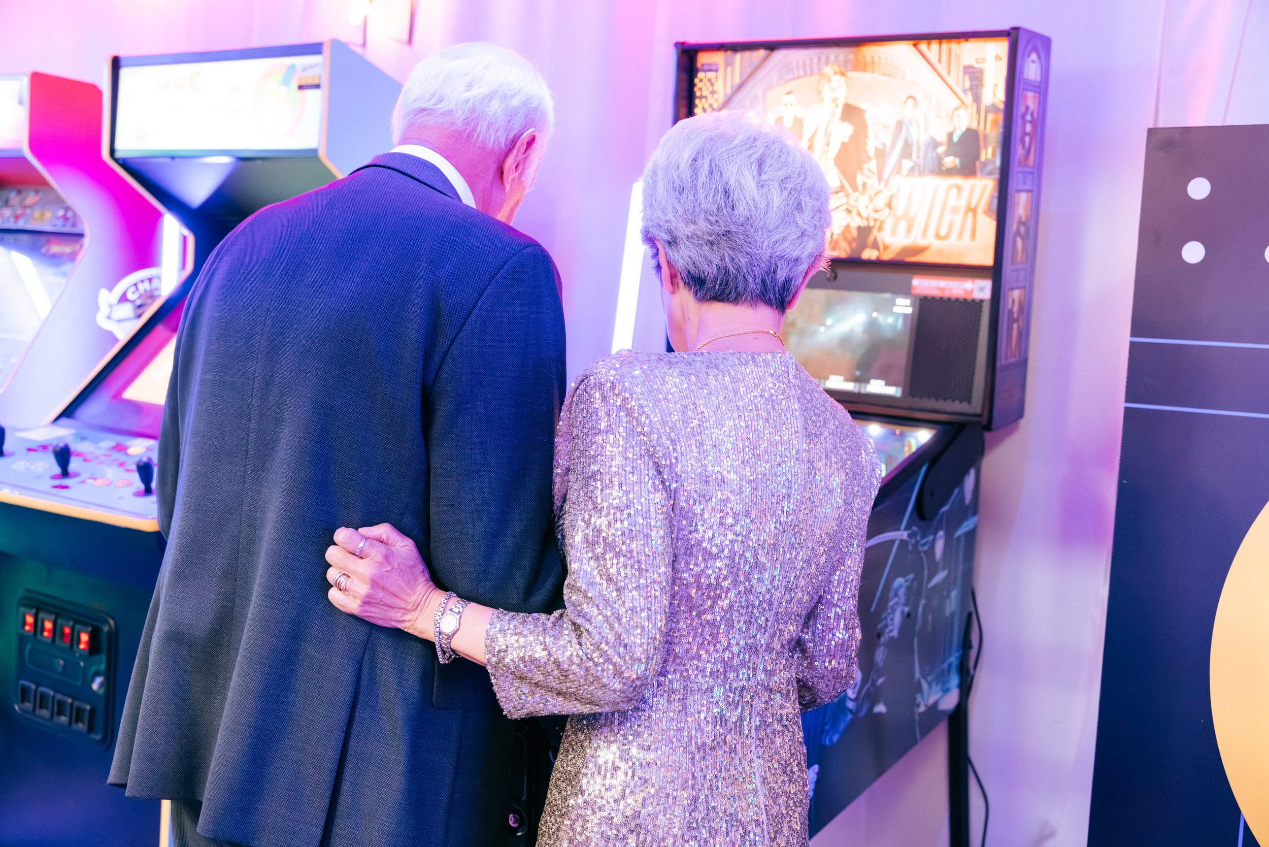 An elderly man and woman, both with gray hair, playing a pinball machine together in an arcade. The woman is wearing a shiny, sequined dress and jewelry, and the man is dressed in a dark suit. The arcade has colorful lighting and other gaming machine