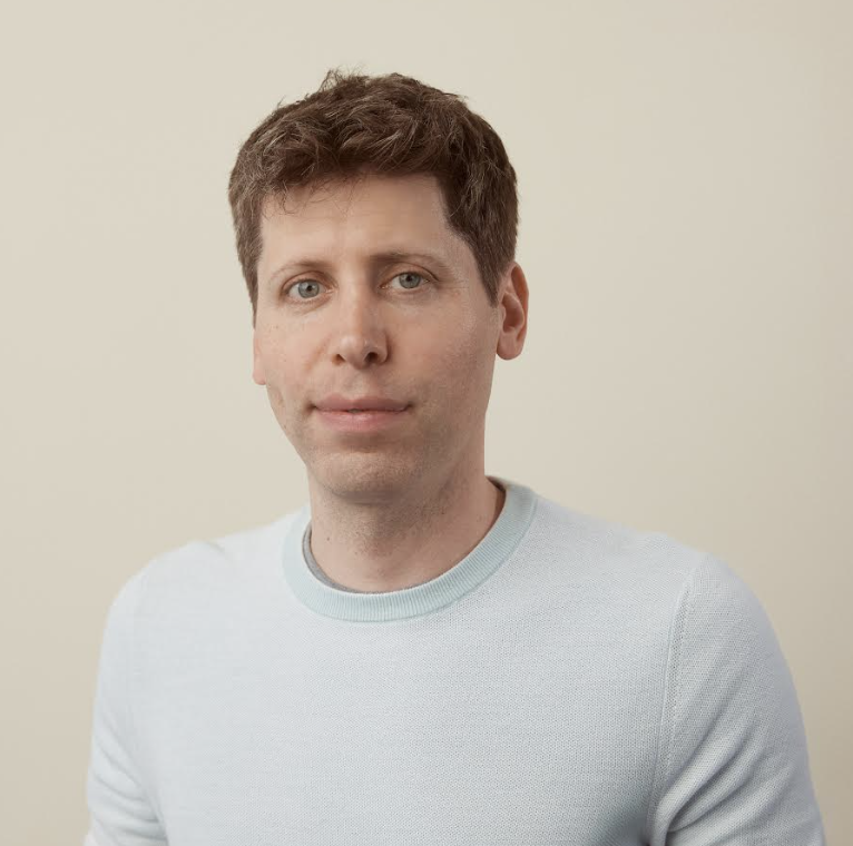 A young man with short brown hair and blue eyes wearing a light gray sweater, standing against a plain off-white background. sam altman