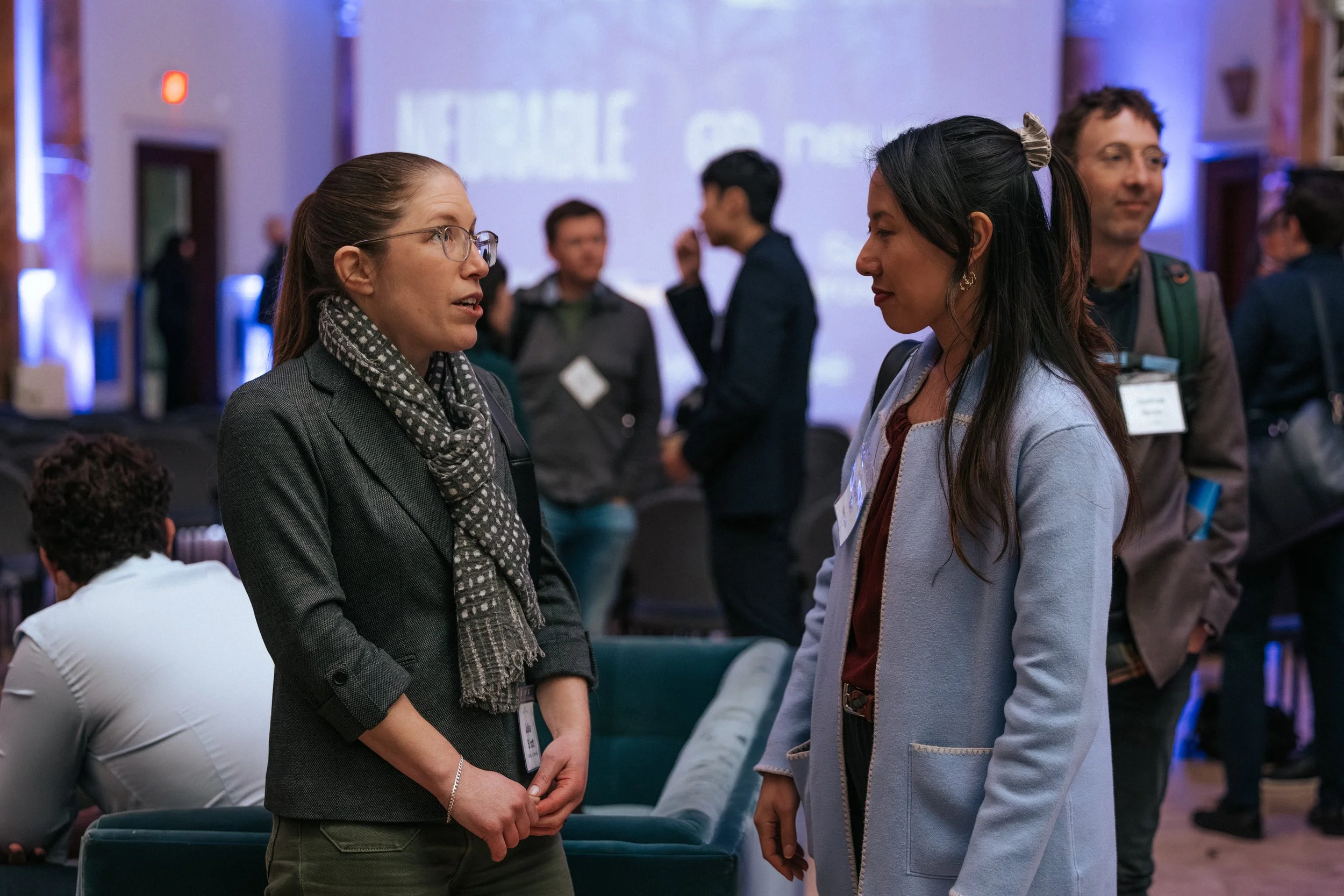 Two women are having a conversation at a professional event or conference. The woman on the left has glasses, a gray blazer, and a polka-dotted scarf. The woman on the right has long dark hair, a light-colored coat, and a name badge. Other people are