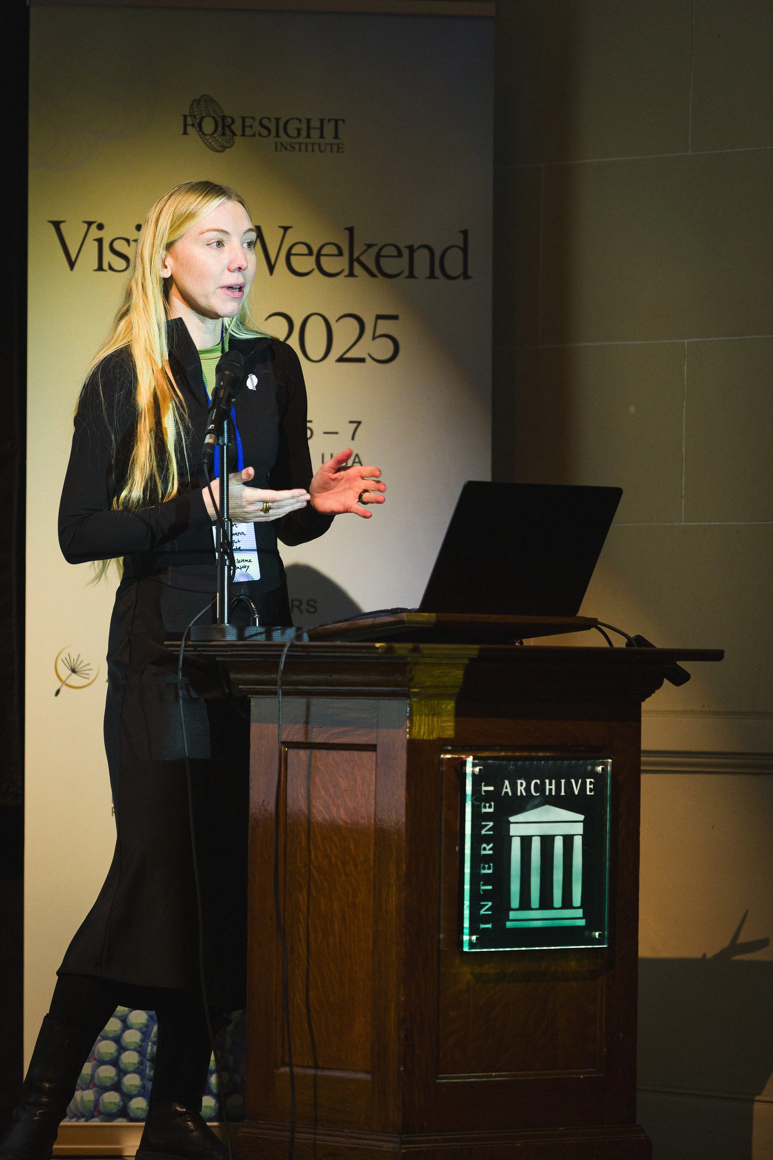 A woman giving a presentation at a conference, standing behind a wooden podium with a laptop, in front of a banner for Foresight Institute's Vision Weekend 2025.