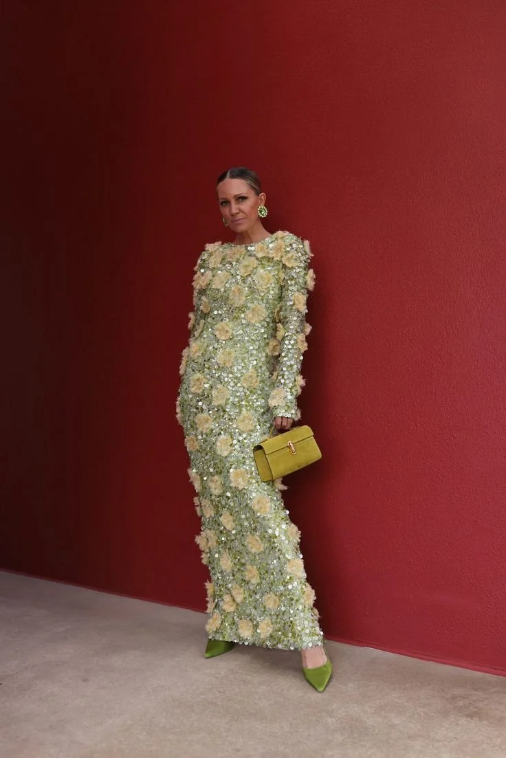 A woman in a long, floral, embellished gown with green shoes, green earrings, and a yellow handbag, standing against a red wall.