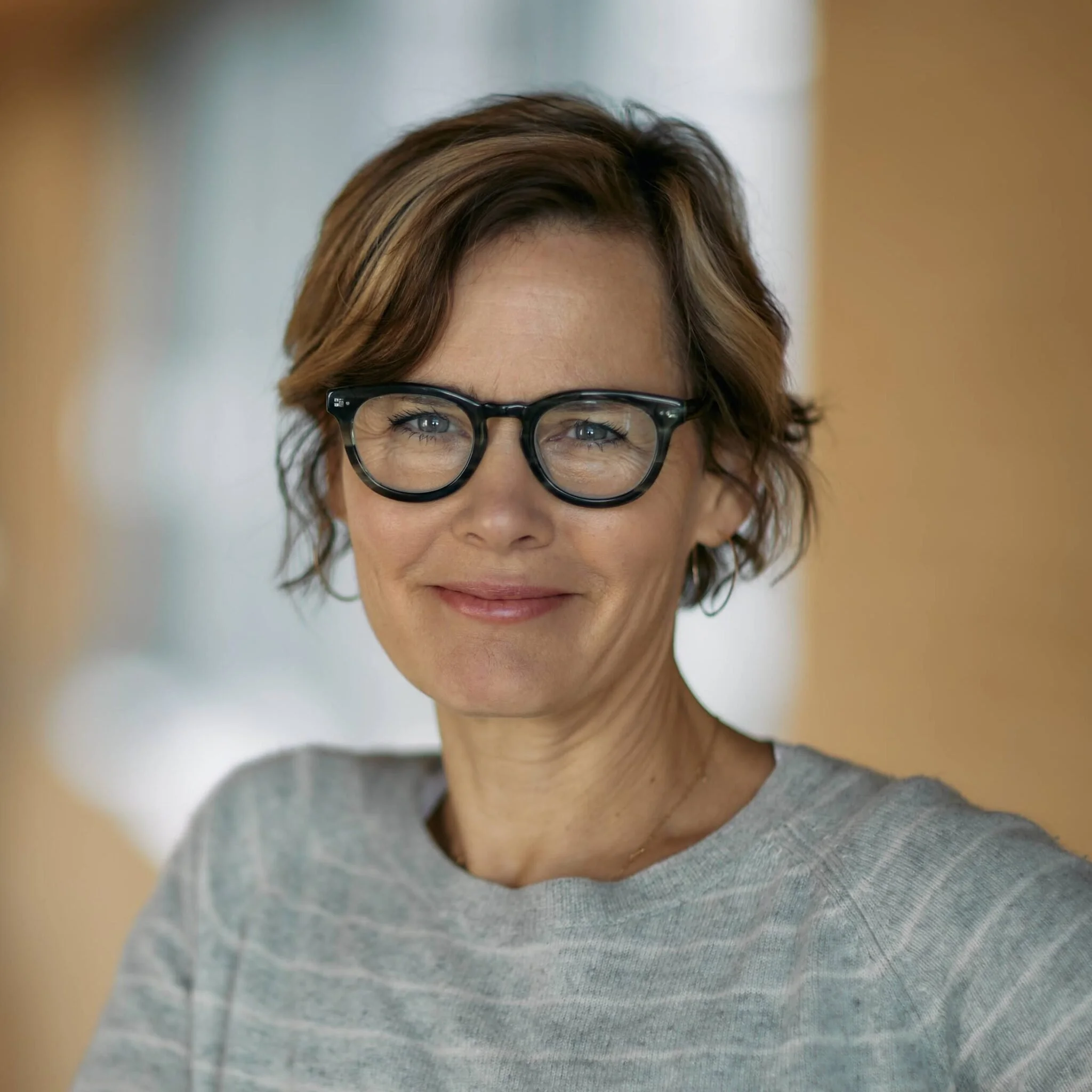 A woman with short, wavy brown hair, wearing black glasses and a light grey sweater, smiling indoors.
