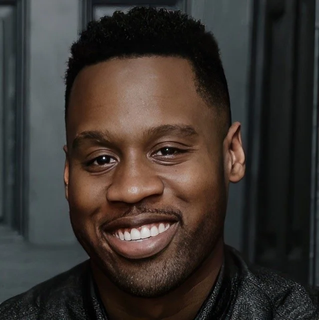 Close-up of smiling man with short curly hair, wearing a black leather jacket, standing in front of a dark background.