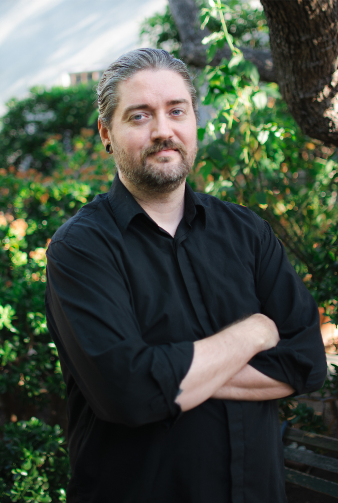 Headshot of Morgan C. Merrill, with gray & brown hair and beard standing outdoors with arms crossed, wearing a black shirt, in a green garden setting.