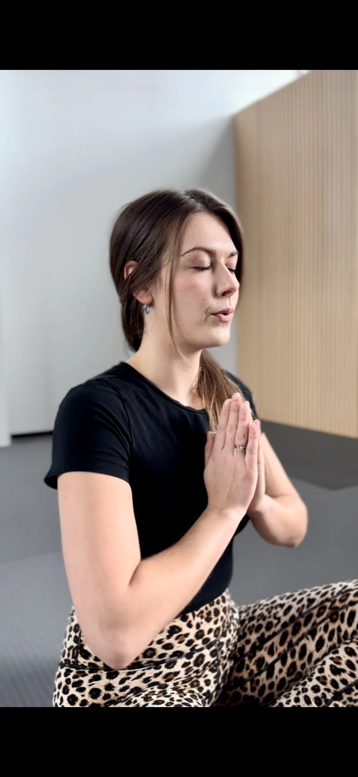 A woman with long brown hair in a ponytail practicing yoga or meditation, with her eyes closed and hands pressed together in a prayer position.