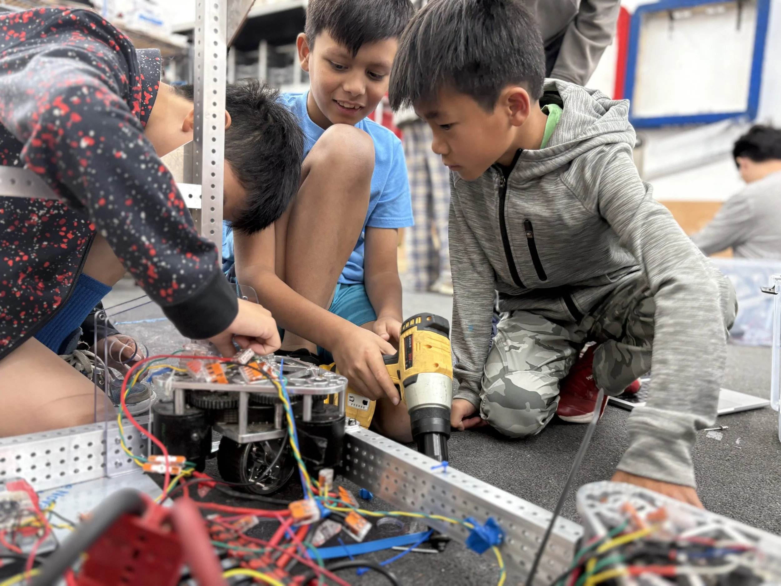 Four boys working together on a robotics project with wires and mechanical parts, some boys are kneeling and using a drill, in a classroom or workshop setting.