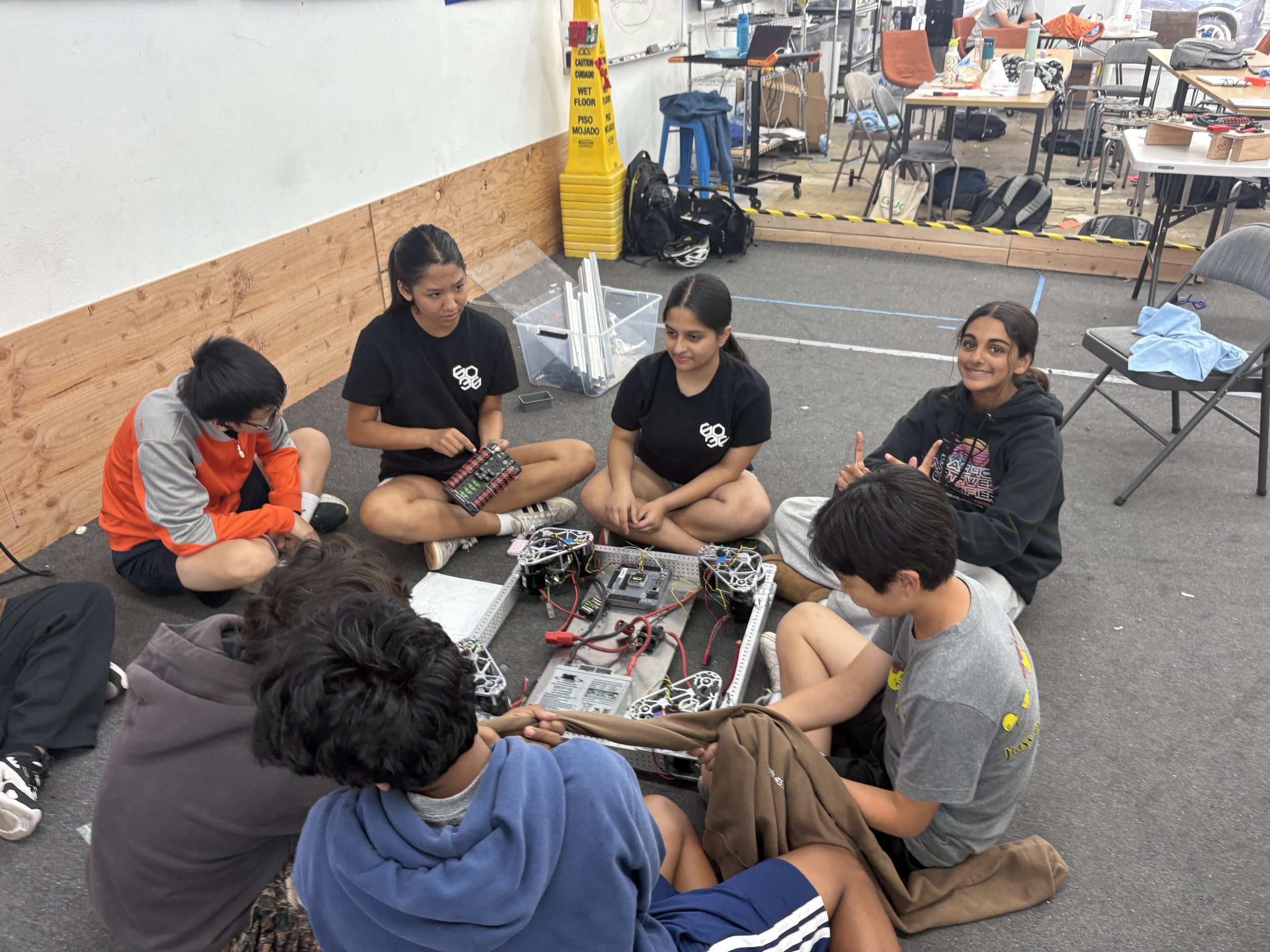 Group of six teenagers sitting cross-legged on the floor in a circle, working on a robotics project with visible wires and robotic parts in the center, in a room with tables, chairs, and various supplies.