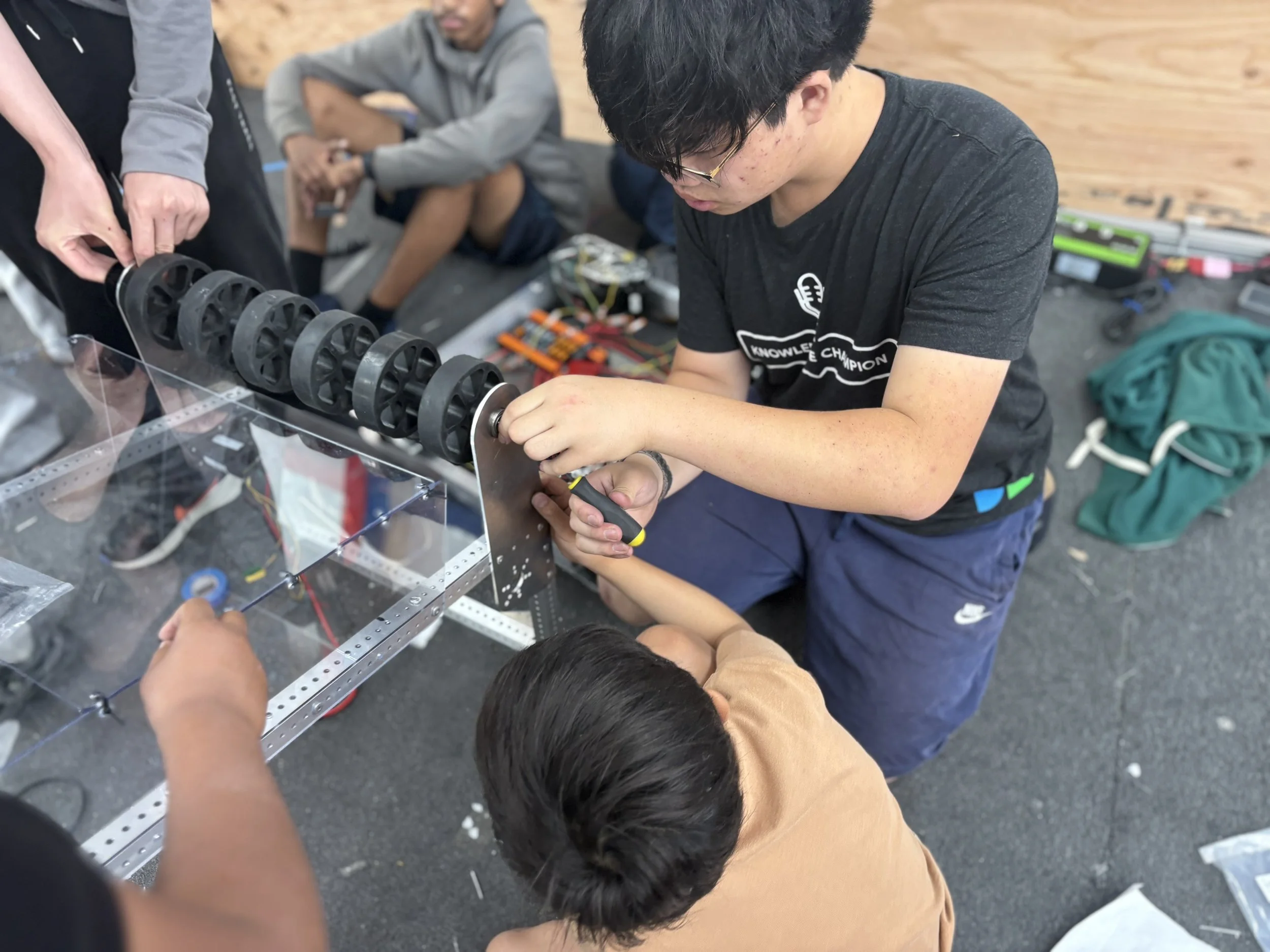 A young person wearing black t-shirt and glasses working on a robotic contraption with a large black wheel assembly, while a child in light brown shirt observes closely. Two other people are standing and sitting nearby, involved in the activity, in a workshop or classroom setting.