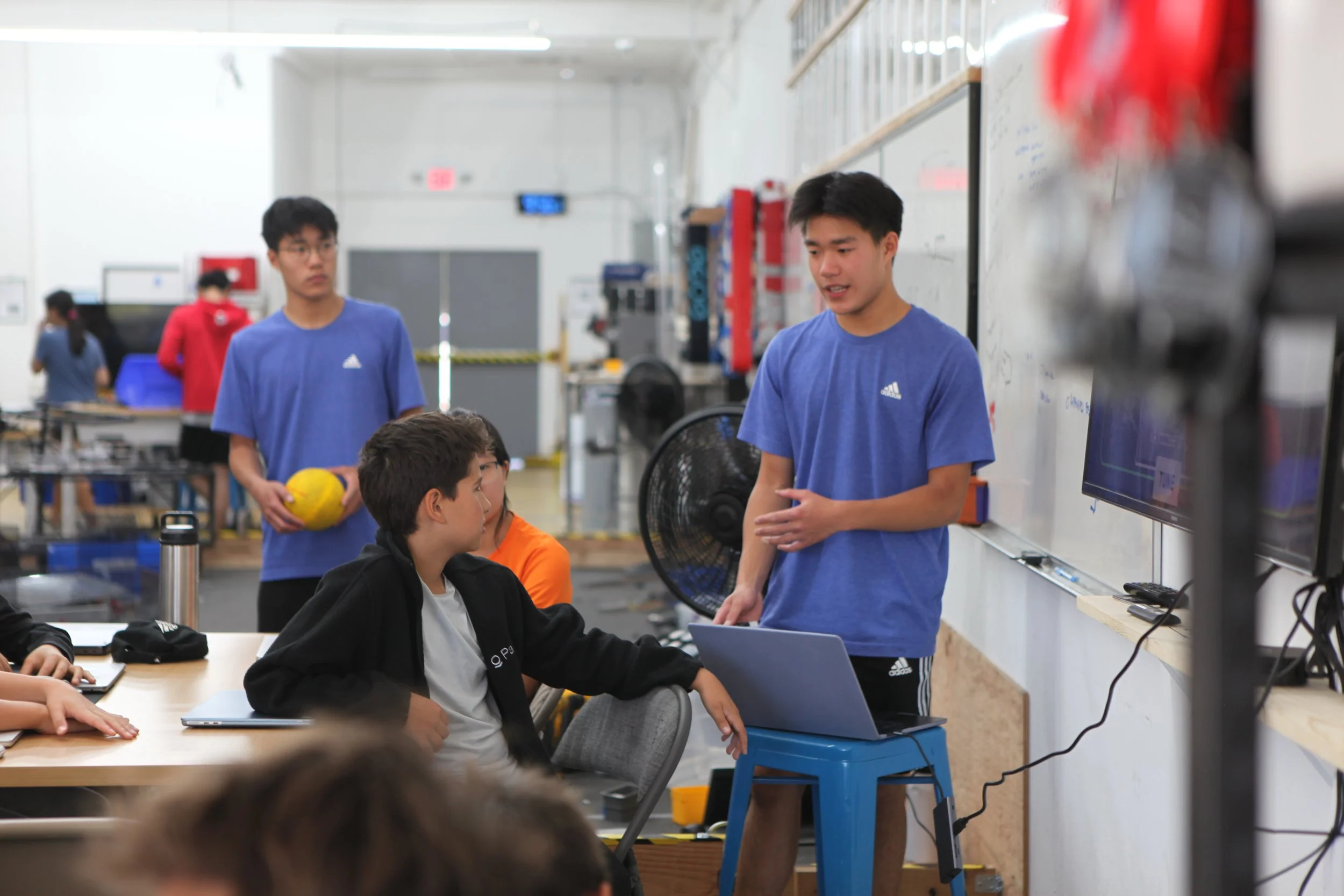 A group of people in a classroom or workshop setting, with one person standing and speaking to seated students. Several students are engaged, some with laptops, and a person in the background holding a yellow ball.