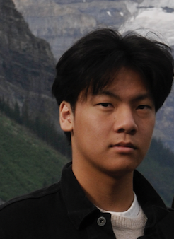 Young man with black hair, wearing a black jacket, standing outdoors with mountains and cloudy sky in the background.