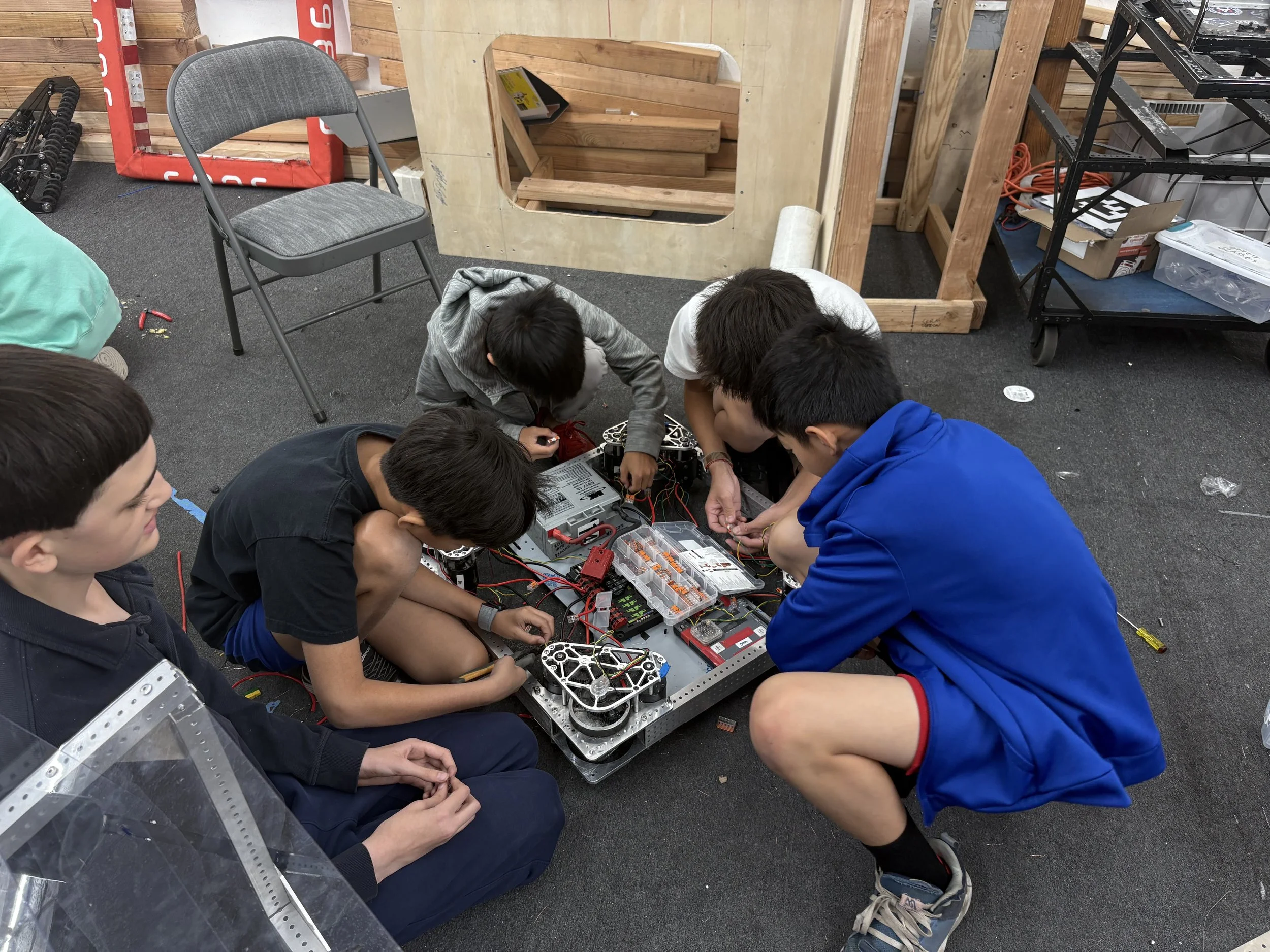 Group of five young students working together on a robotics project on the floor of a workshop or classroom, surrounded by tools and equipment.