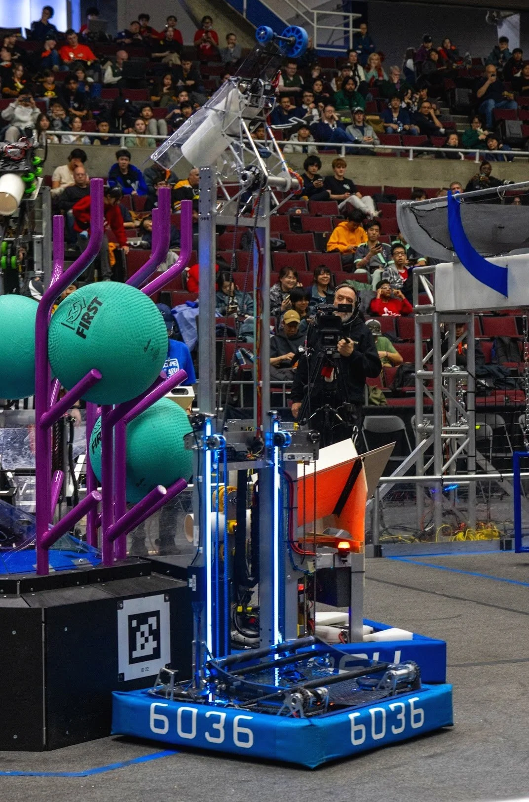 A robotics competition with a robot on the field, surrounded by spectators in the stands. The robot has teal balls, purple bars, and various mechanical components. An operator is controlling it, with a cameraman nearby.