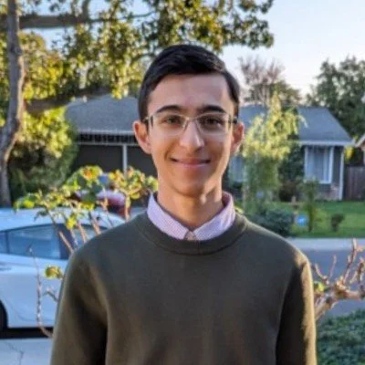 A young man with glasses smiling outdoors in a residential neighborhood with trees and houses in the background.