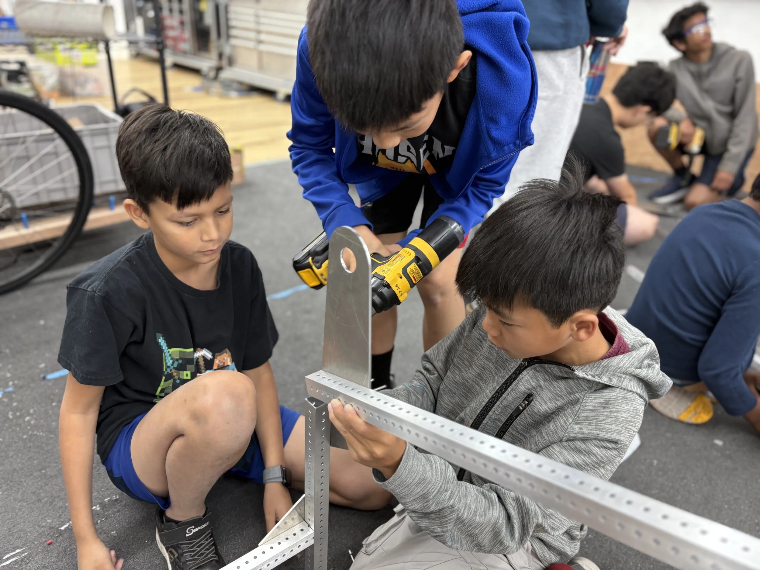 Children working on a robotics project; two kids kneeling and one standing, using a power drill on a metal frame.