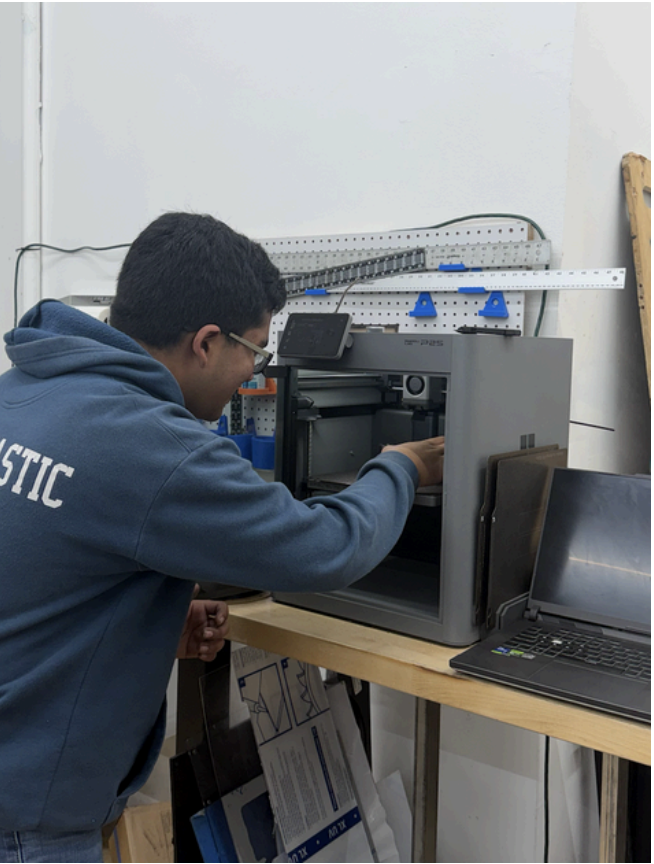 A young man in a blue hoodie with 'FUTURISTIC' printed on the sleeve is working on a 3D printer at a wooden table. There is a laptop next to the printer, and a pegboard with tools and wires on the white wall behind.