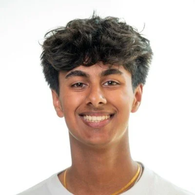 Young man with curly dark hair and a bright smile, wearing a white shirt and a gold chain, against a white background.