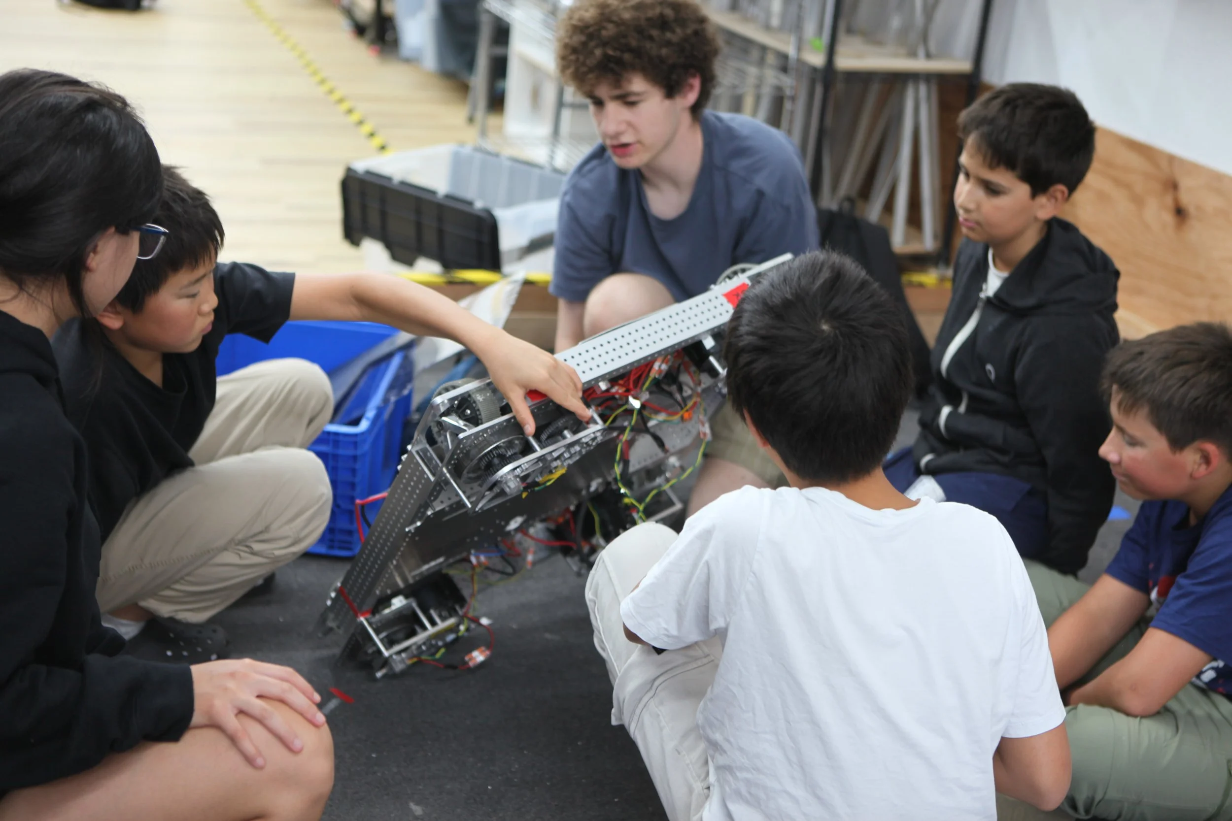 Group of children and a young adult working on a robot or electronic project, gathered around it and discussing.