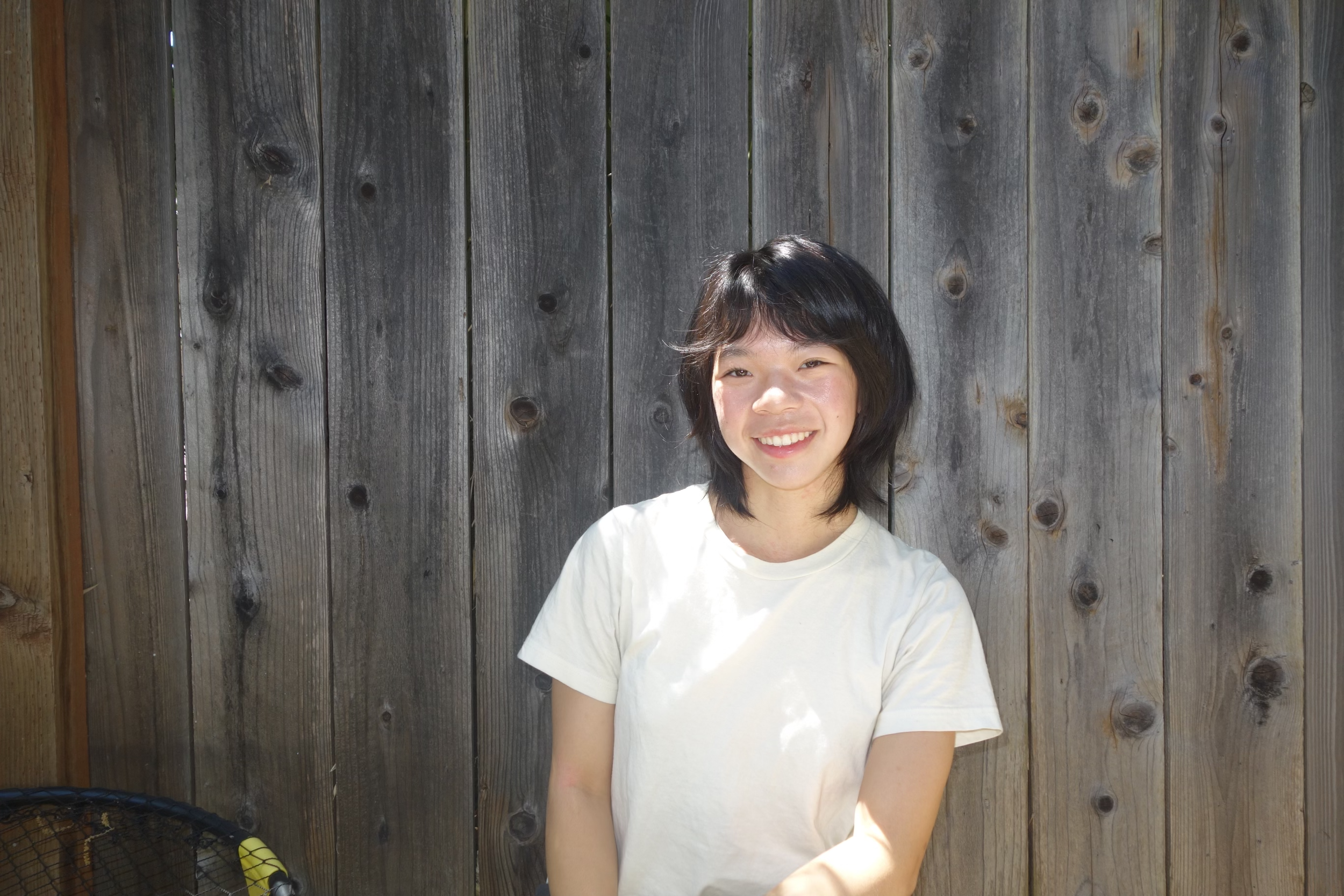 A young woman with black hair and a white T-shirt smiling in front of a wooden fence.