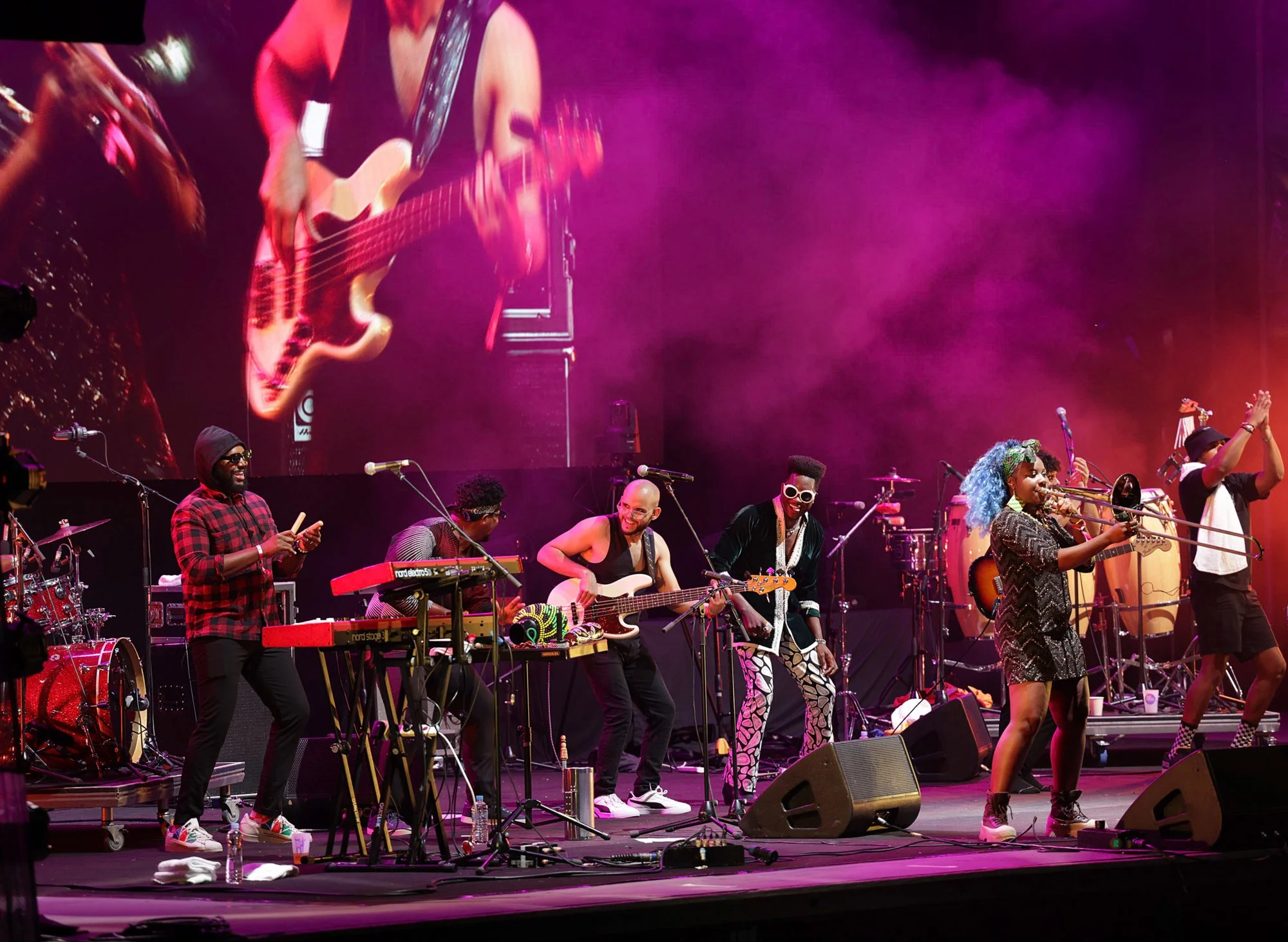 A live musical performance on stage featuring multiple musicians playing instruments such as guitar, keyboard, drums, and trumpet. The stage is lit with vibrant pink and purple lighting, and performers are dressed in colorful and casual attire, with 