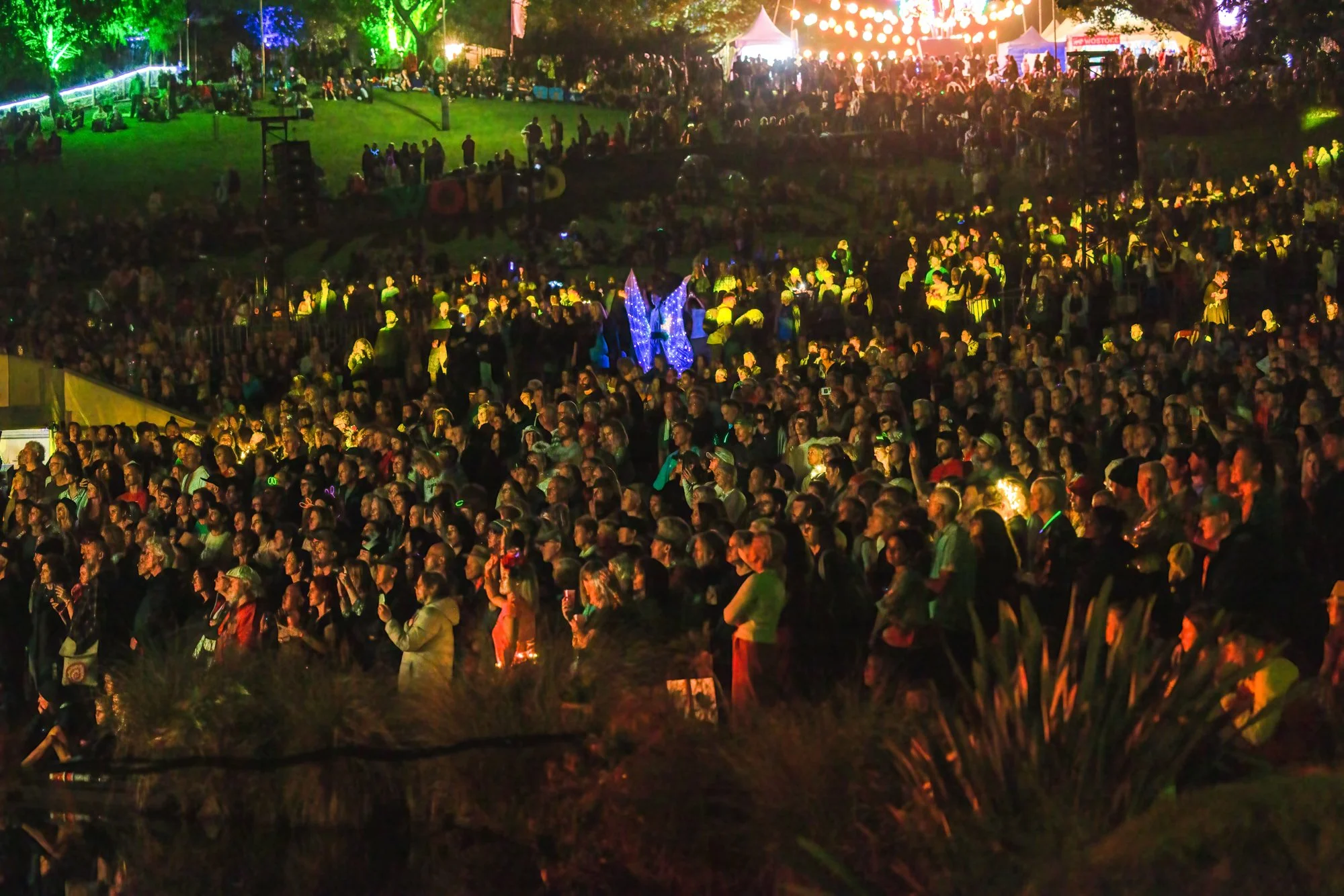 A large crowd at an outdoor nighttime event with colorful lights, food tents, and a stage in the background.