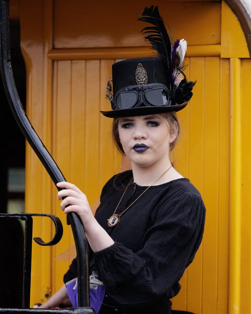 Young woman in gothic fashion with black lipstick, wearing a black top hat adorned with feathers and goggles, standing in front of a yellow train car.