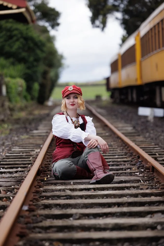 A young woman sitting on railway tracks next to a yellow train, wearing a red cap, white blouse, red vest, green pants, and red boots, with trees and cloudy sky in the background.