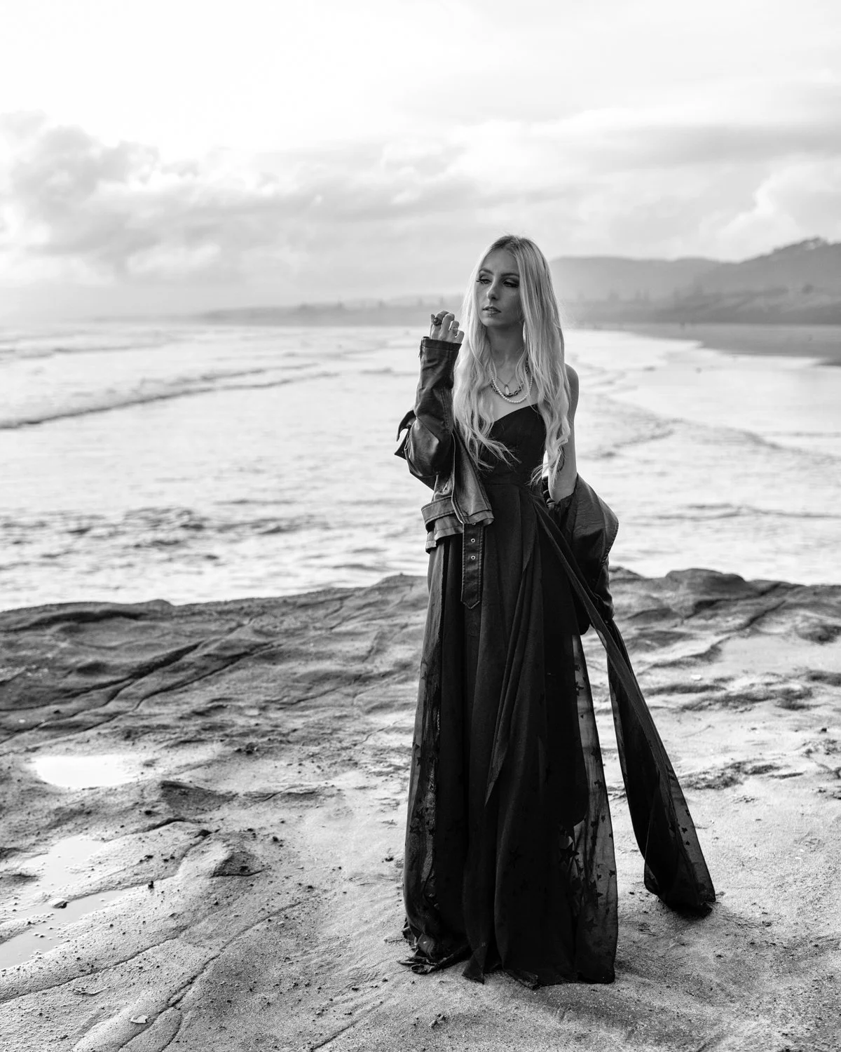 A woman with long blonde hair wearing a black dress and a leather jacket standing on a rocky beach with waves and distant hills in the background, in black and white.