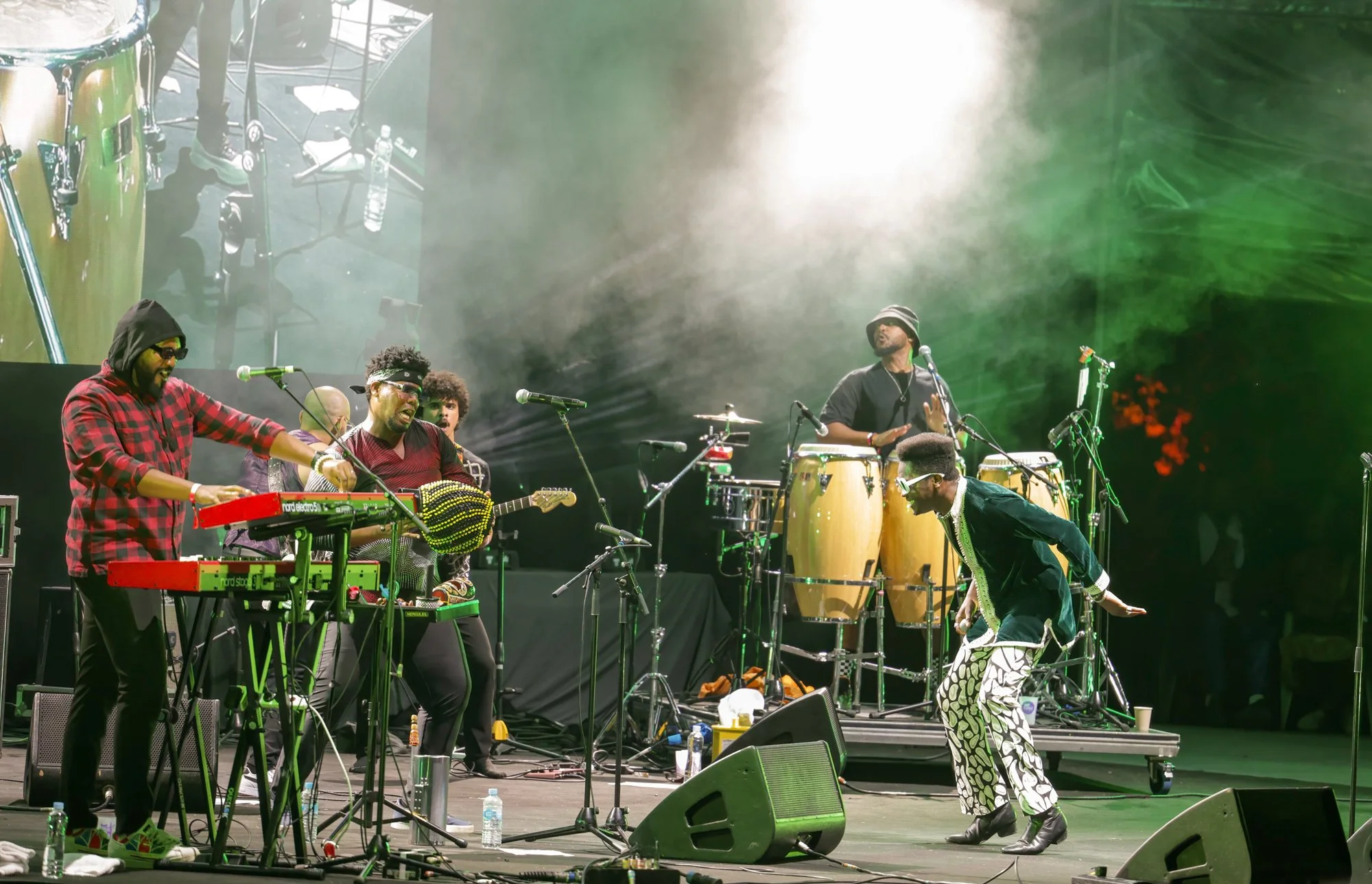 A music band performing on stage with multiple musicians playing instruments and a singer in patterned pants. Bright stage lights and smoke effects enhance the lively atmosphere.