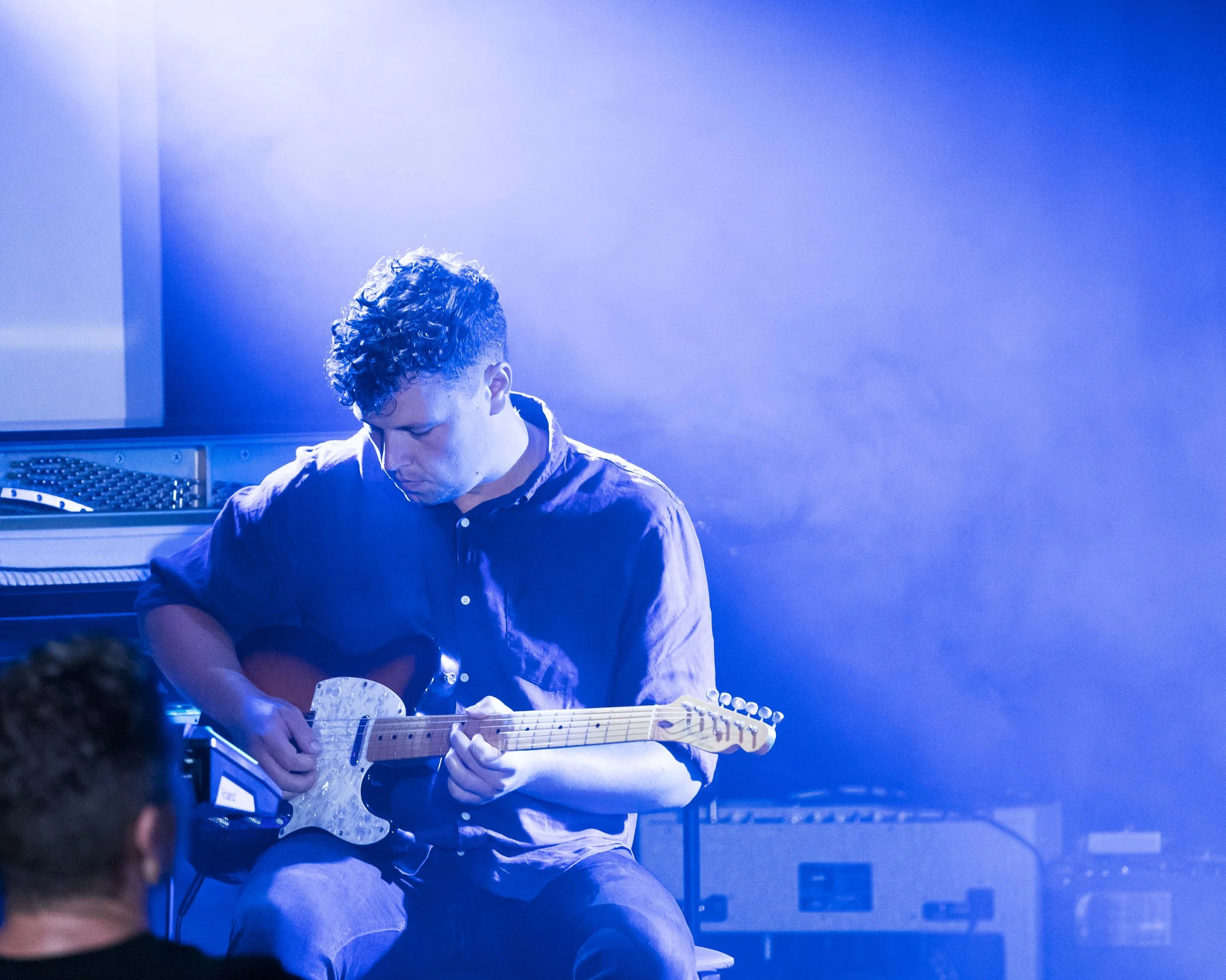 A person with curly hair playing an electric guitar in a blue-lit room.