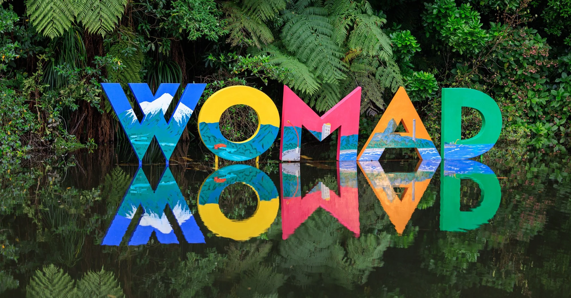 Colorful letters spelling 'WONDER' reflect on a body of water, with lush green fern and plant foliage in the background.