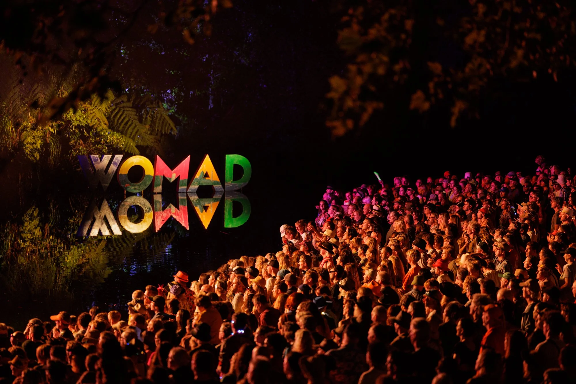 Night scene of a large crowd gathered by a river, with colorful decorations including illuminated 'WOMAD' sign and trees reflected in the water.