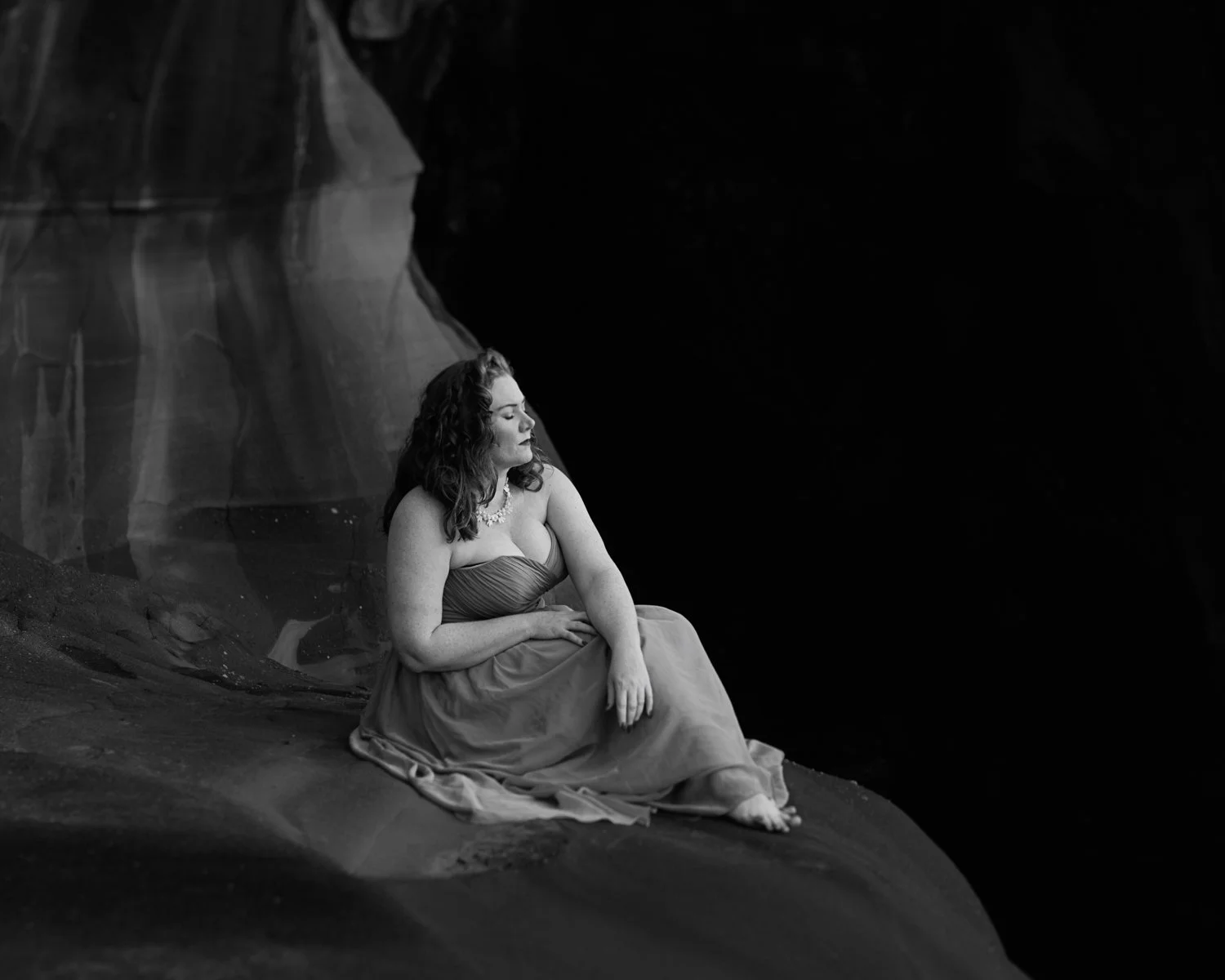 A woman sitting on a rock near water, eyes closed, in a black and white photograph.
