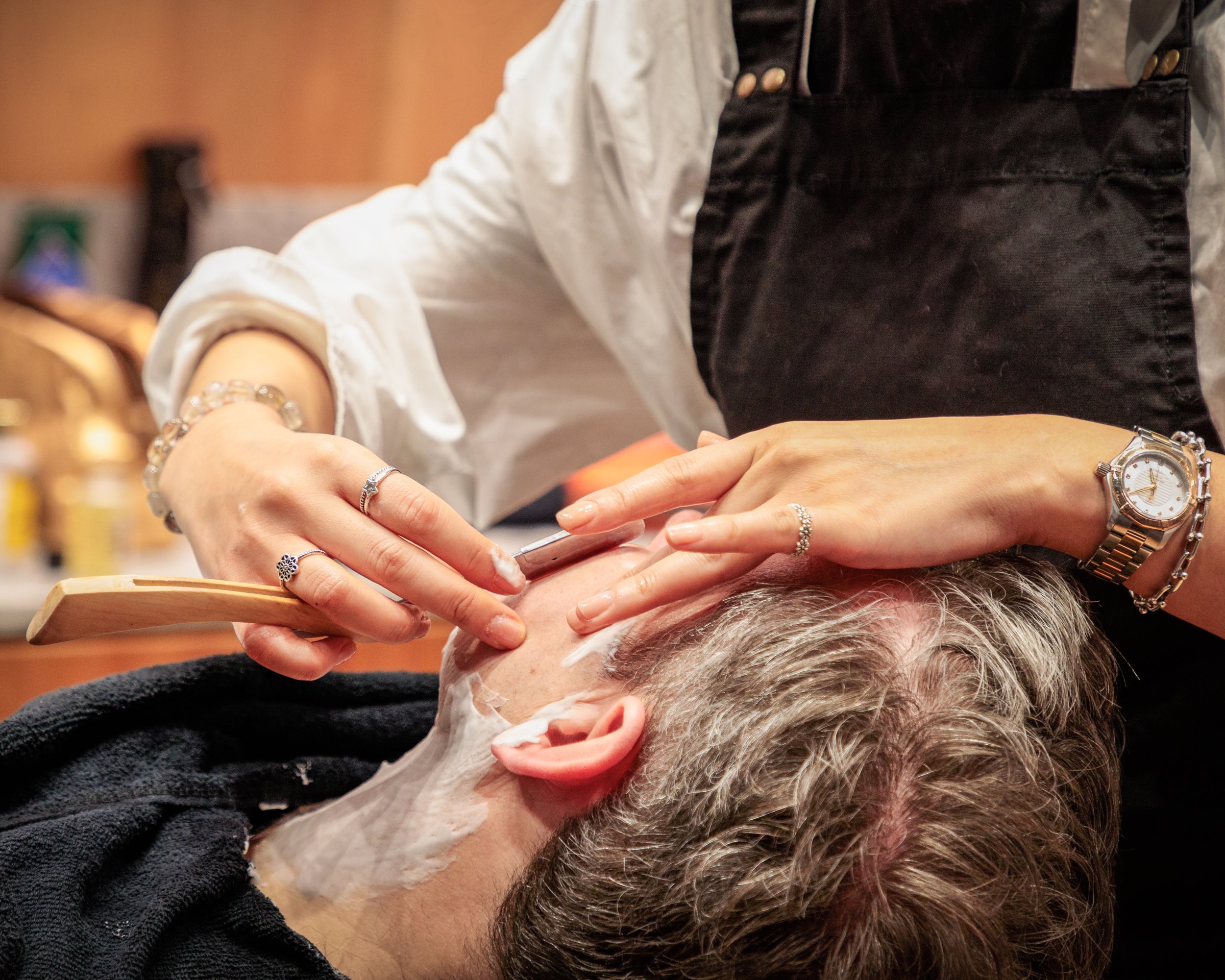A person giving a haircut to an older individual with grey hair at a salon. The person's hands are visible, holding a comb and scissors, and they are wearing rings and a watch. The person appears to be focusing on trimming the hair.