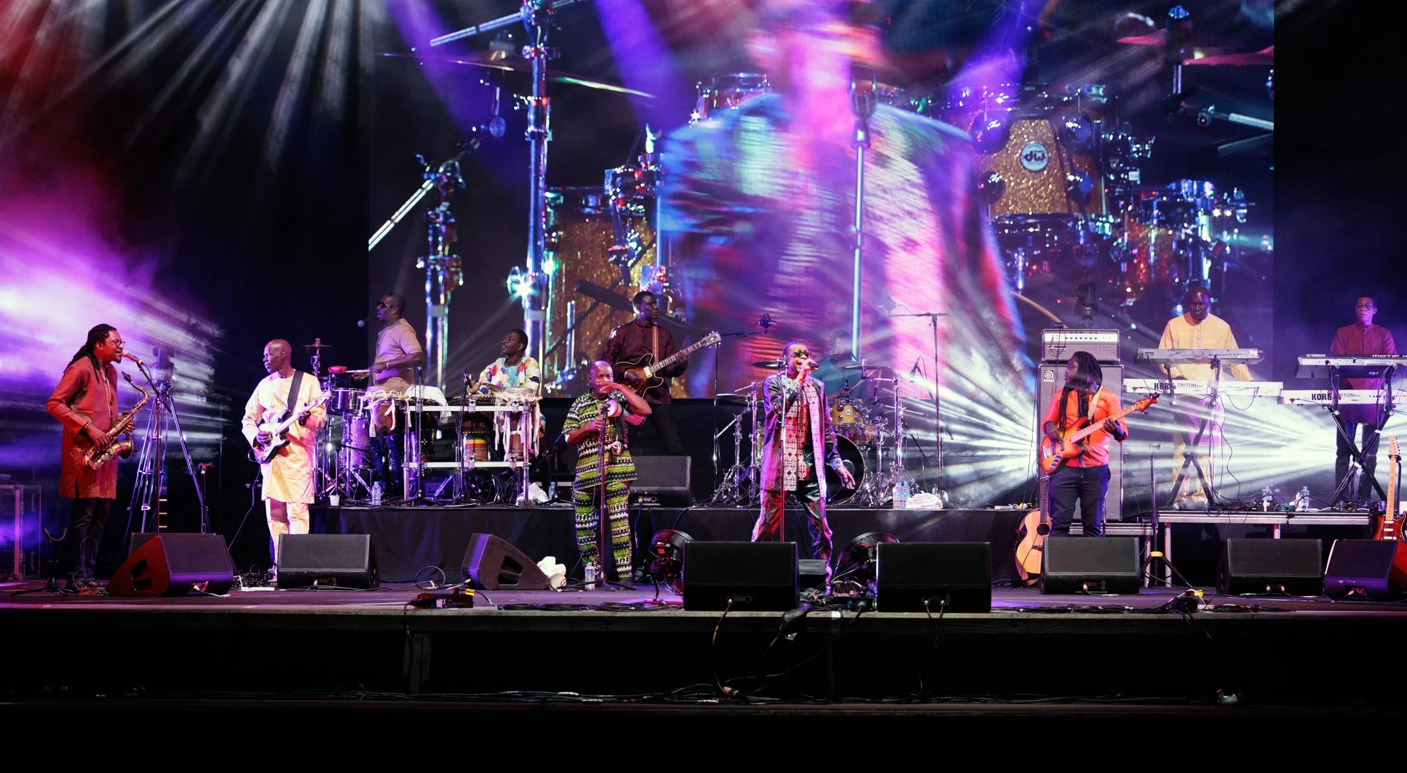 African musicians performing on stage with colorful lights and a large screen in the background, playing various instruments including guitars, keyboards, and drums.