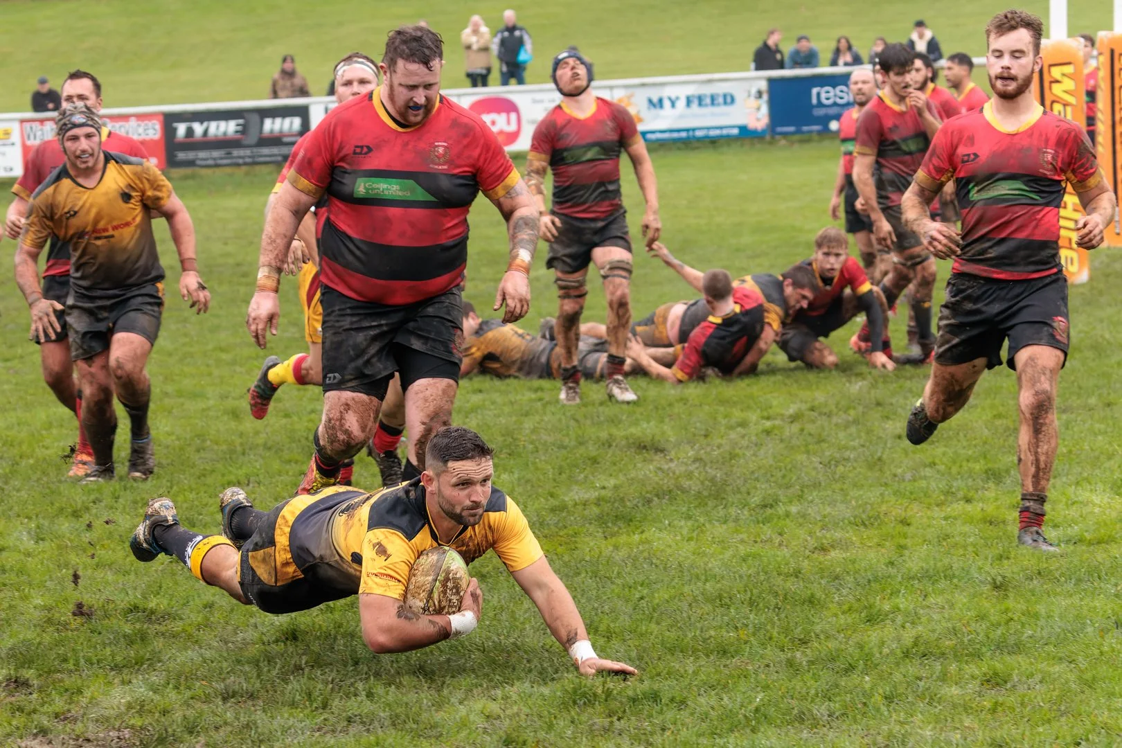 Rugby players in muddy uniforms on a grassy field during a match, with some players on the ground and others running or standing.