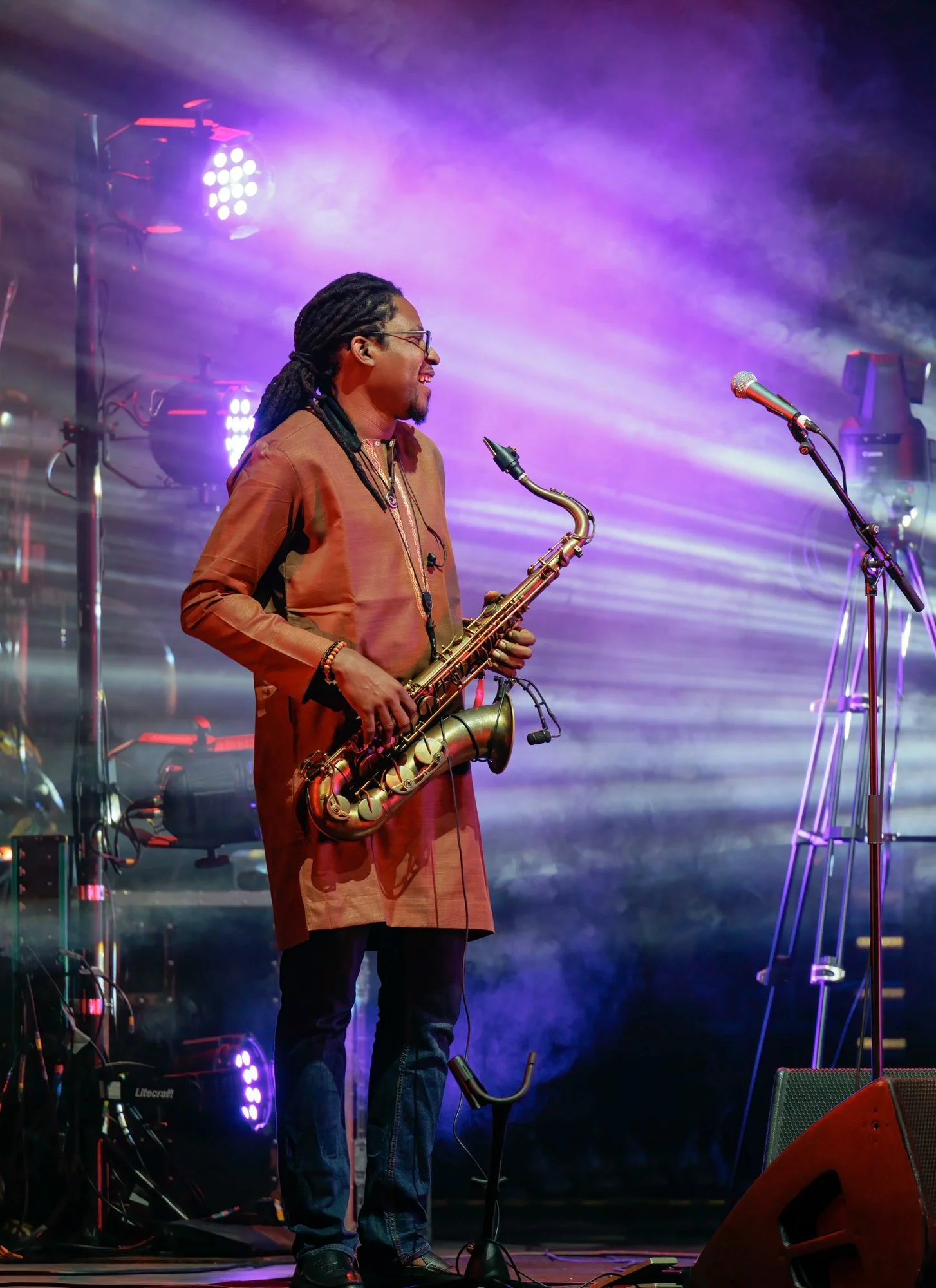 A man with dreadlocks and glasses playing an electric keyboard on stage with colorful purple and white stage lights and fogging effect.