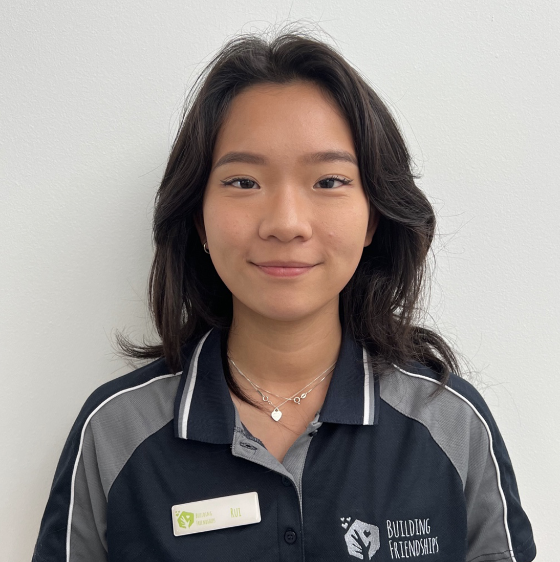 A woman with blonde hair smiling, wearing a dark jacket with patches, standing in front of a beige textured wall.