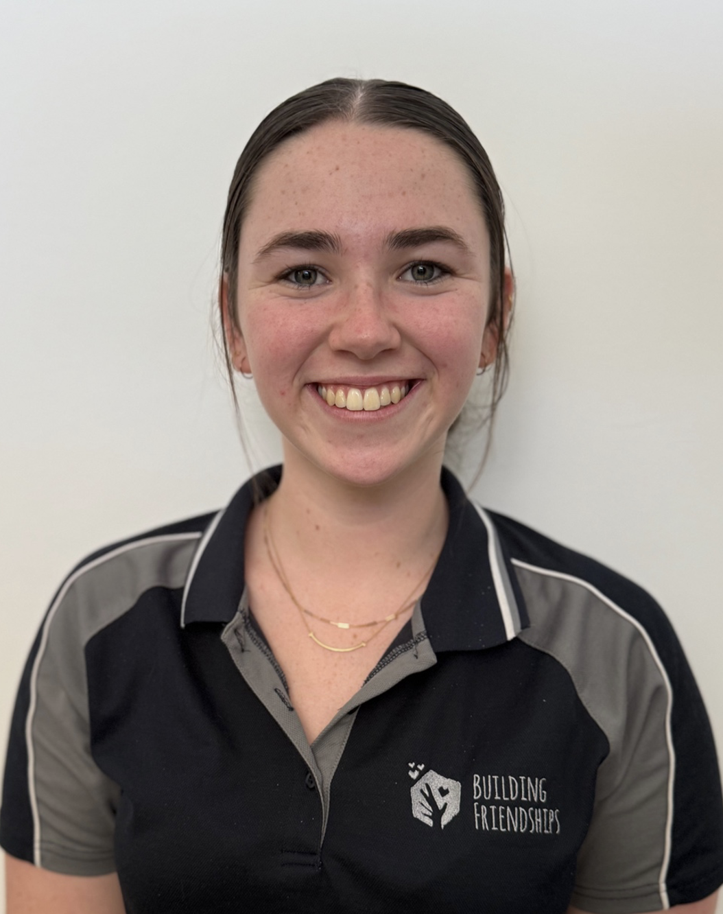 A smiling young woman with blonde hair wearing a black and gray polo shirt with the words 'BUILDING FRIENDSHIPS' and a logo on it, standing against a beige wall.