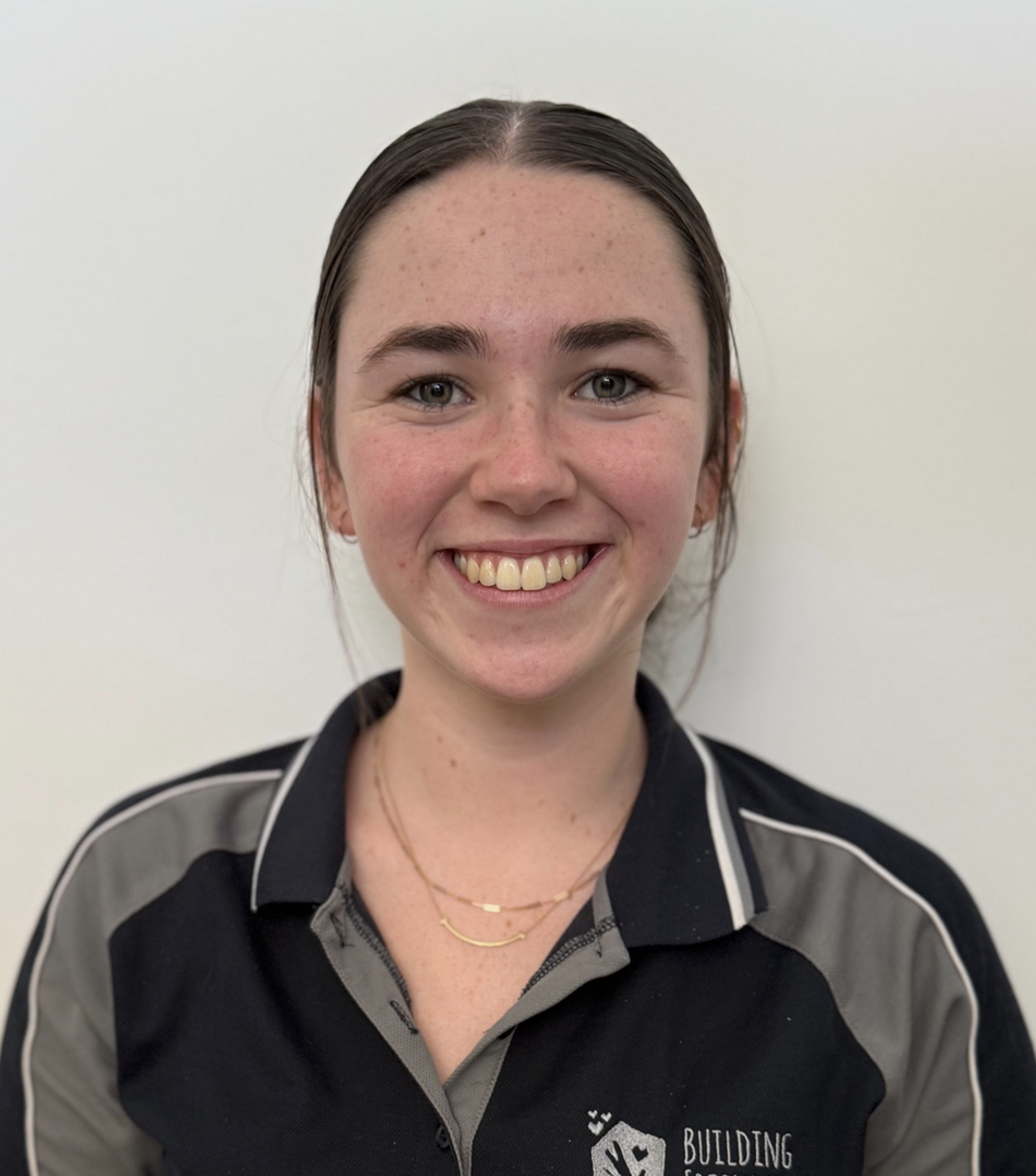 A smiling young woman with blonde hair wearing a black and gray polo shirt with the words 'BUILDING FRIENDSHIPS' and a logo on it, standing against a beige wall.