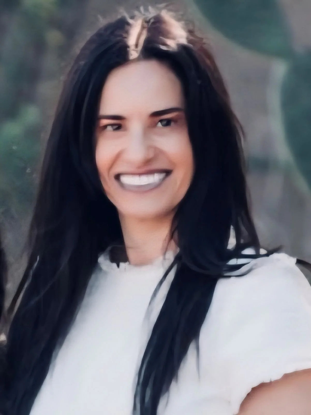 A Jewish Woman with dark hair, smiling, with her chin resting on her hand, wearing a beige jacket and a ring on her finger, indoors near a wooden partition and window.