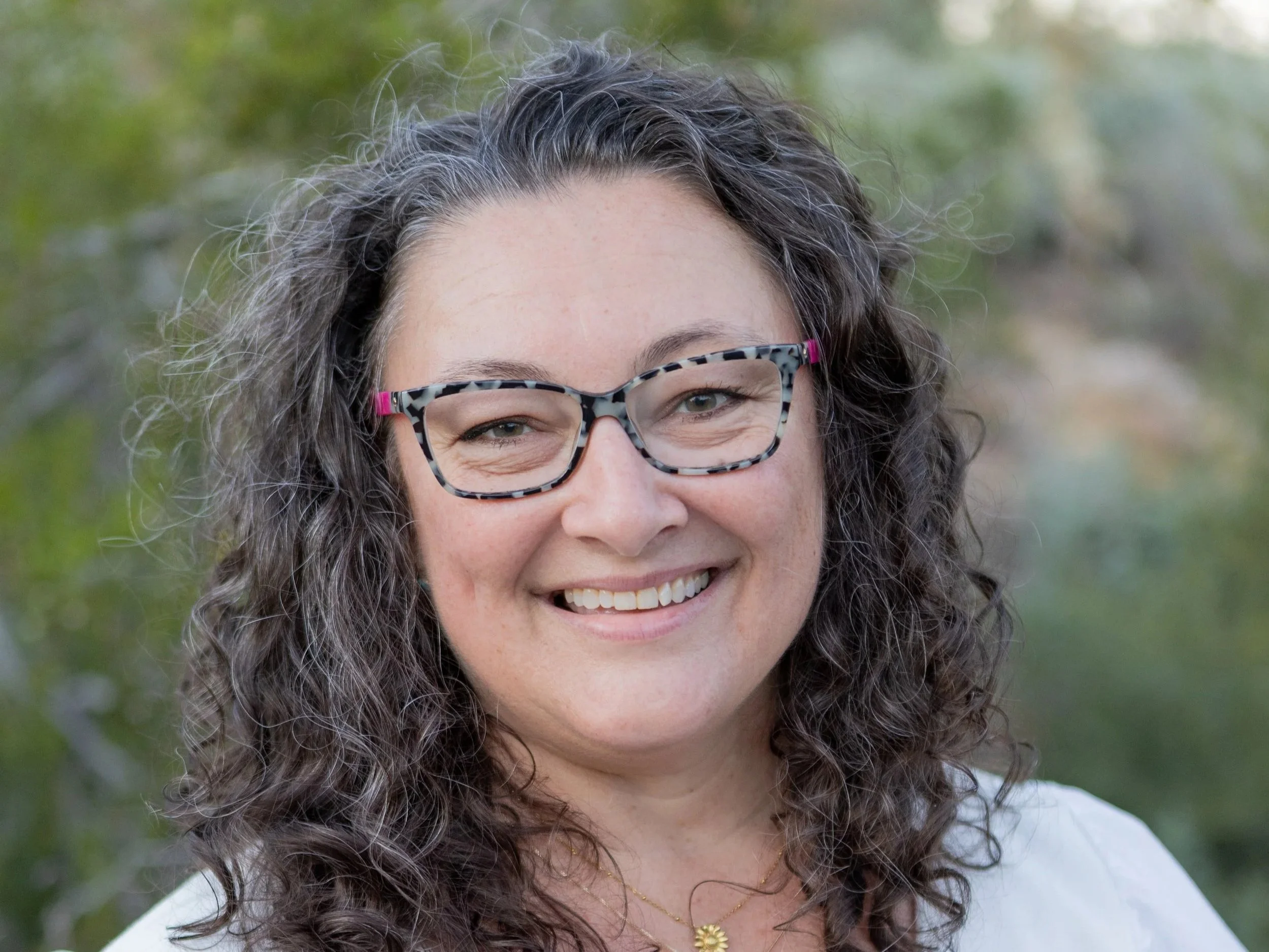 A woman with curly dark hair wearing glasses and a blue knitted sweater smiling in front of a beige textured wall.
