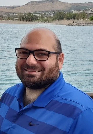 Portrait of a middle eastern man with glasses, a beard, and short dark hair, wearing a blue plaid shirt, against a black background.