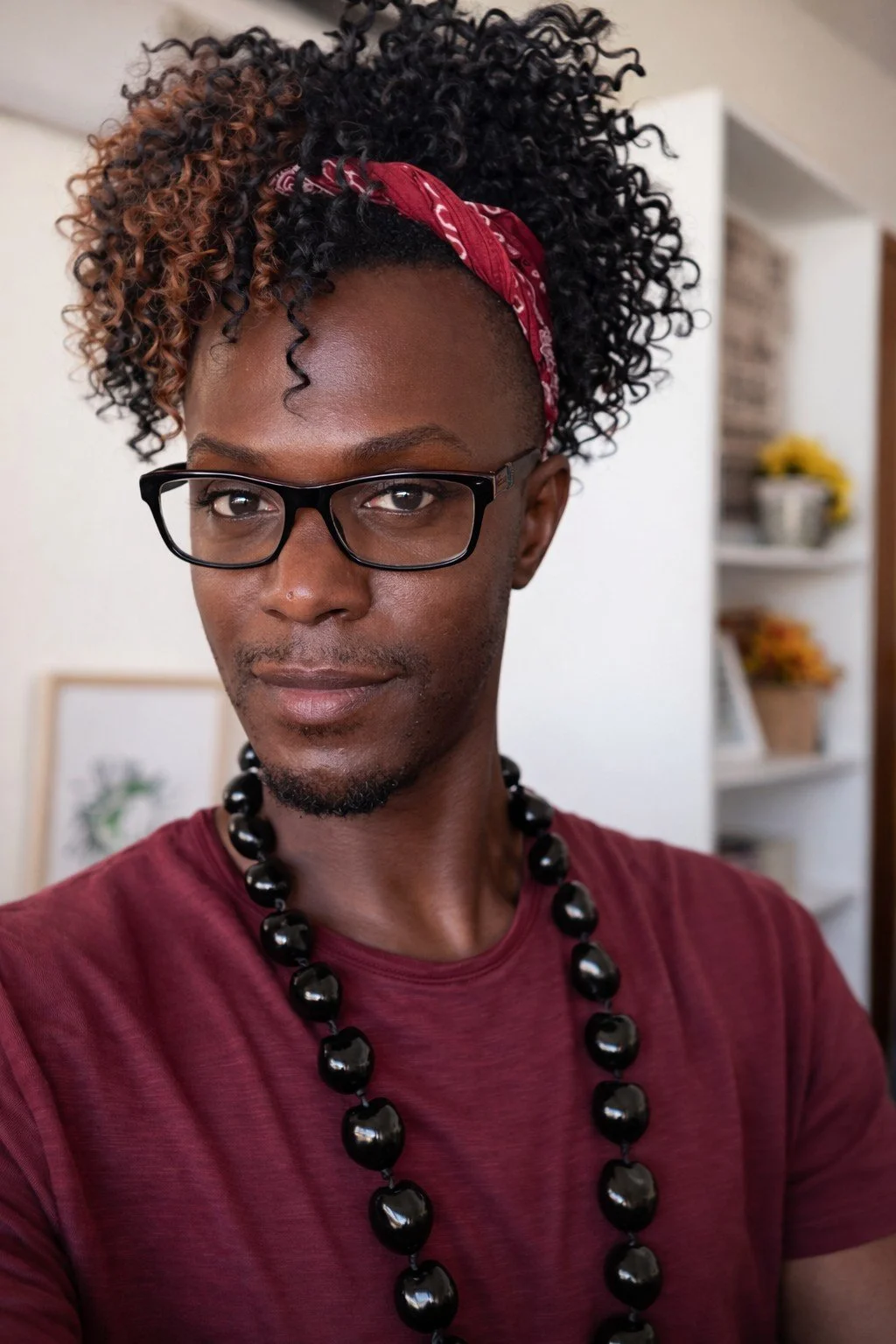 An African American male with curly black and brown hair, wearing glasses, a red bandana, a maroon shirt, and a black beaded necklace, standing indoors.