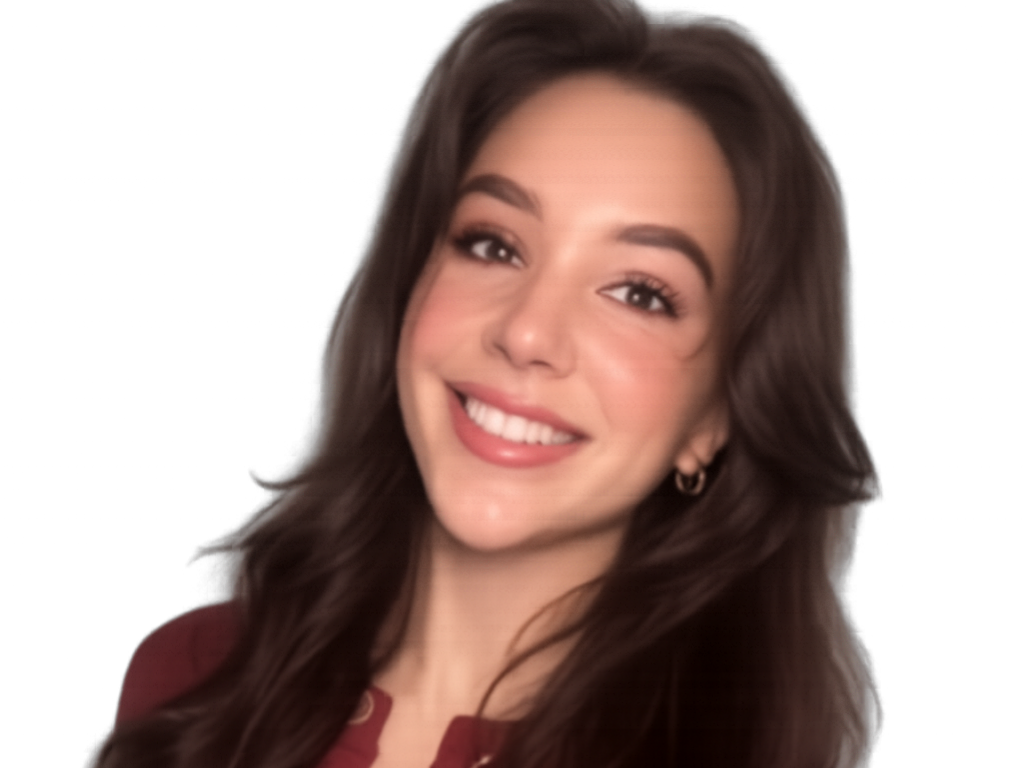 A young woman with long, wavy brown hair smiling at the camera, wearing a maroon button-up top and gold jewelry, standing against a plain wall.