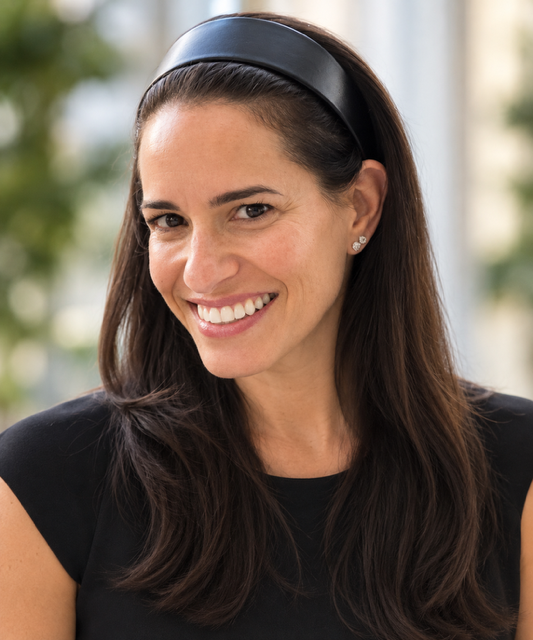 A Jewish Woman with dark hair, smiling, with her chin resting on her hand, wearing a beige jacket and a ring on her finger, indoors near a wooden partition and window.