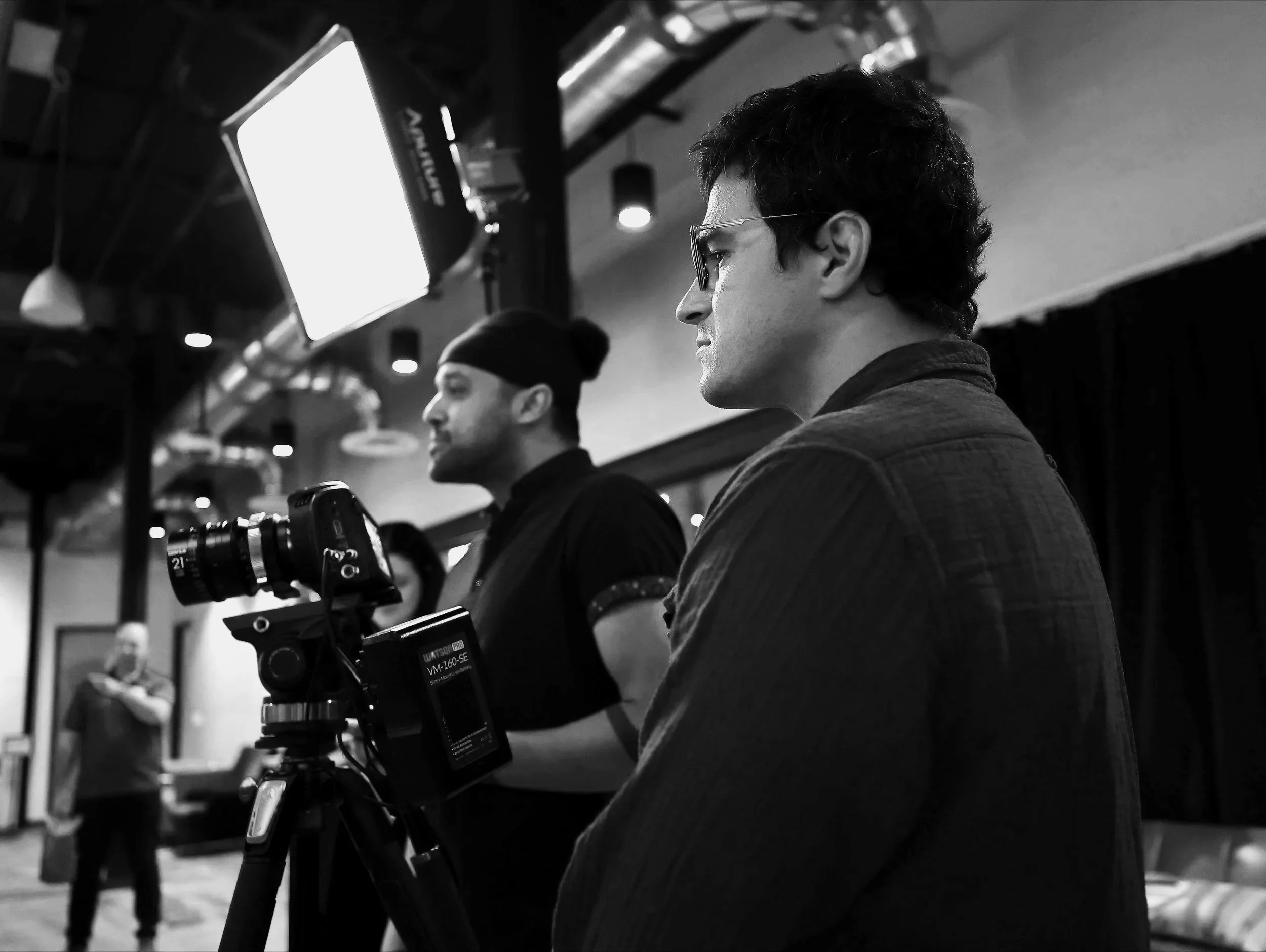 A man with curly hair and glasses operating a camera on a tripod in a studio setting, with another person in the background.