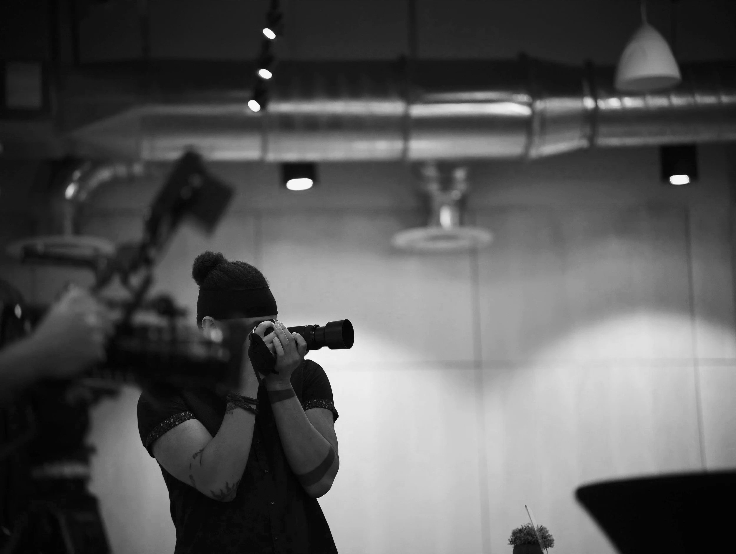 A person wearing a beanie and holding a camera to their face, shooting a black and white photograph in an indoor studio with hanging lights and industrial piping.