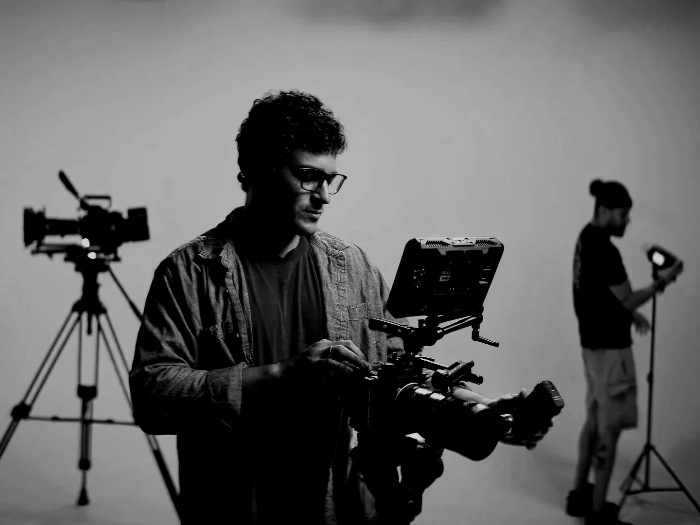 Black and white photo of a man operating a professional camera on a tripod in a studio. In the background, a woman is adjusting lighting equipment.
