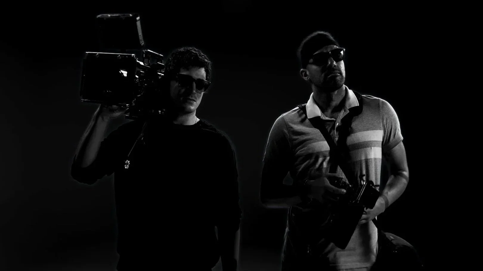 Two men with sunglasses holding cameras, standing in a dark setting, one with a shoulder-mounted camera and the other with a handheld camera.