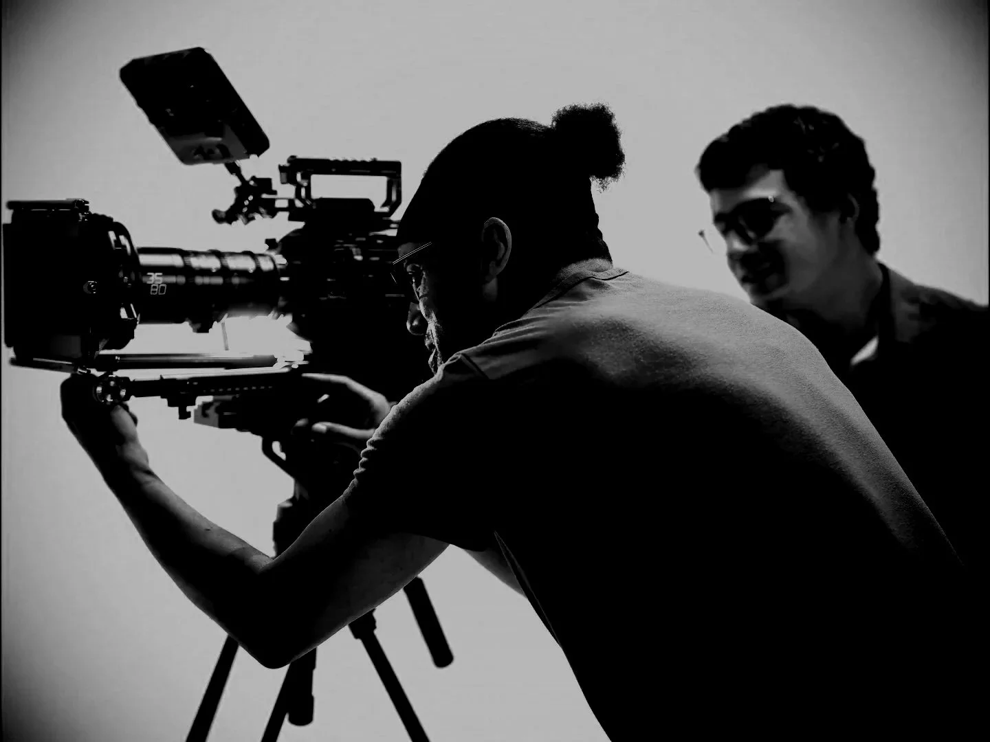 Two men working with a camera on a tripod in a studio, one looking through the camera and the other standing behind, both in profile view.
